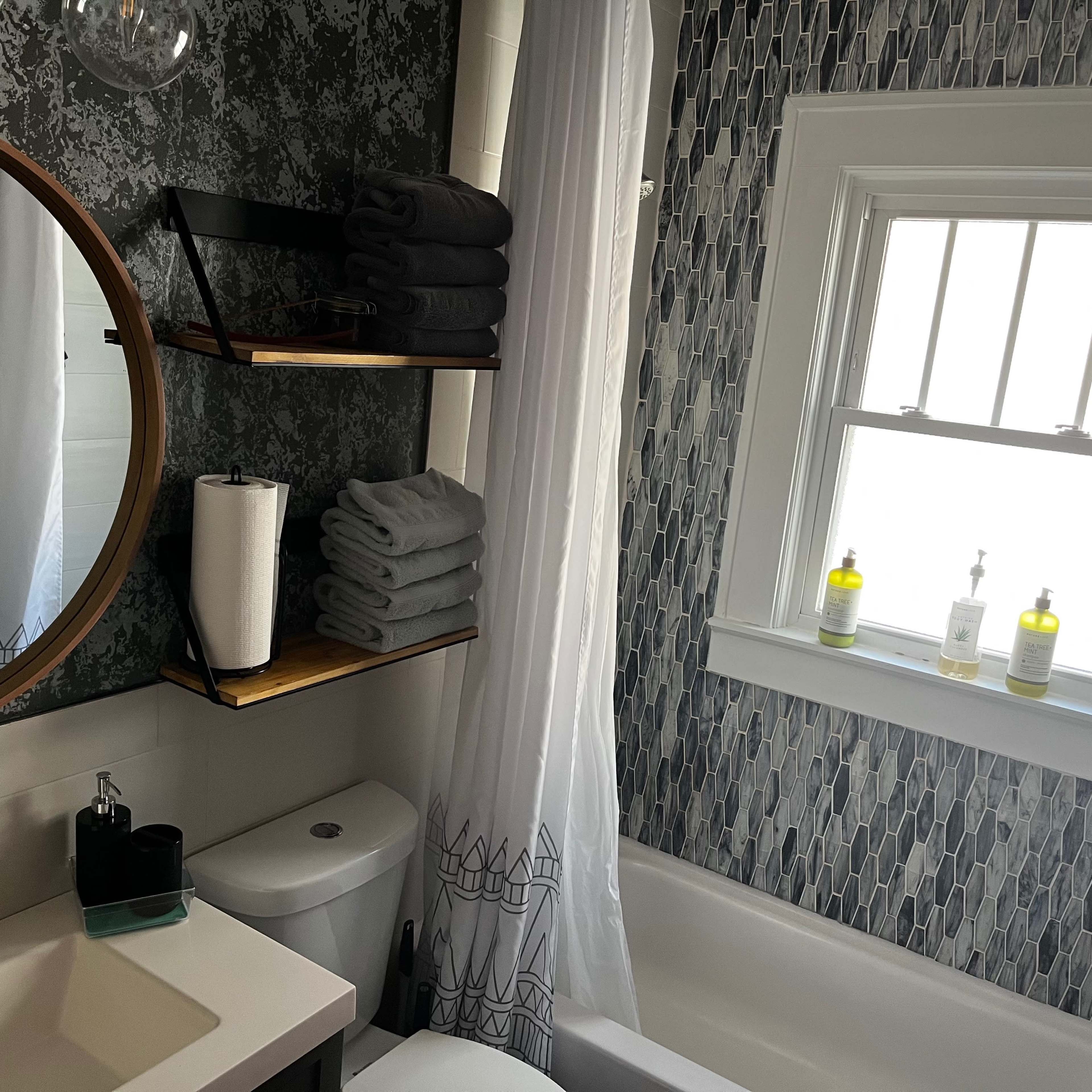 A modern bathroom featuring a round mirror, a bathtub, and shelves with neatly stacked towels and toiletries beside a window.