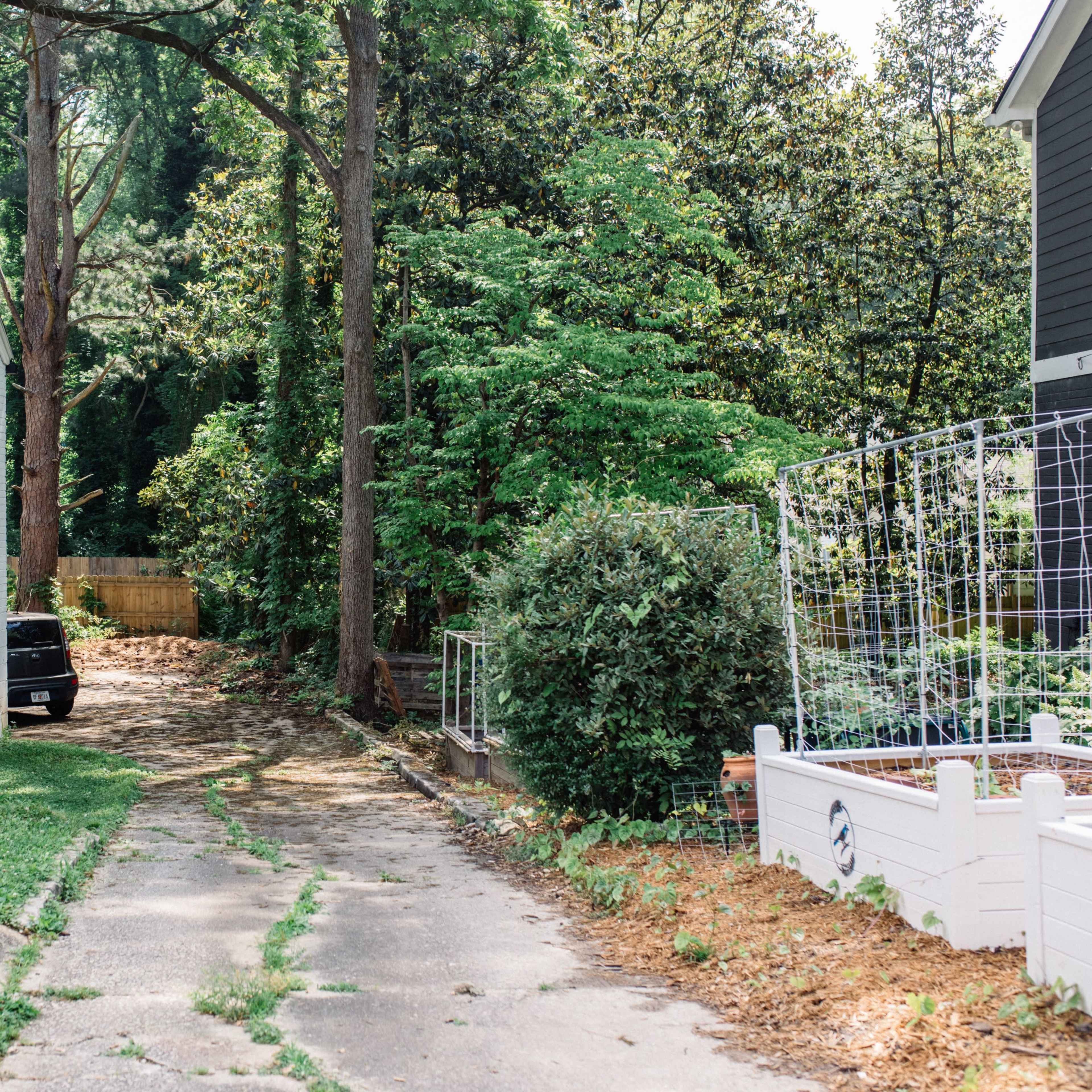 A gravel driveway runs alongside a white brick house and leads towards a wooded area.