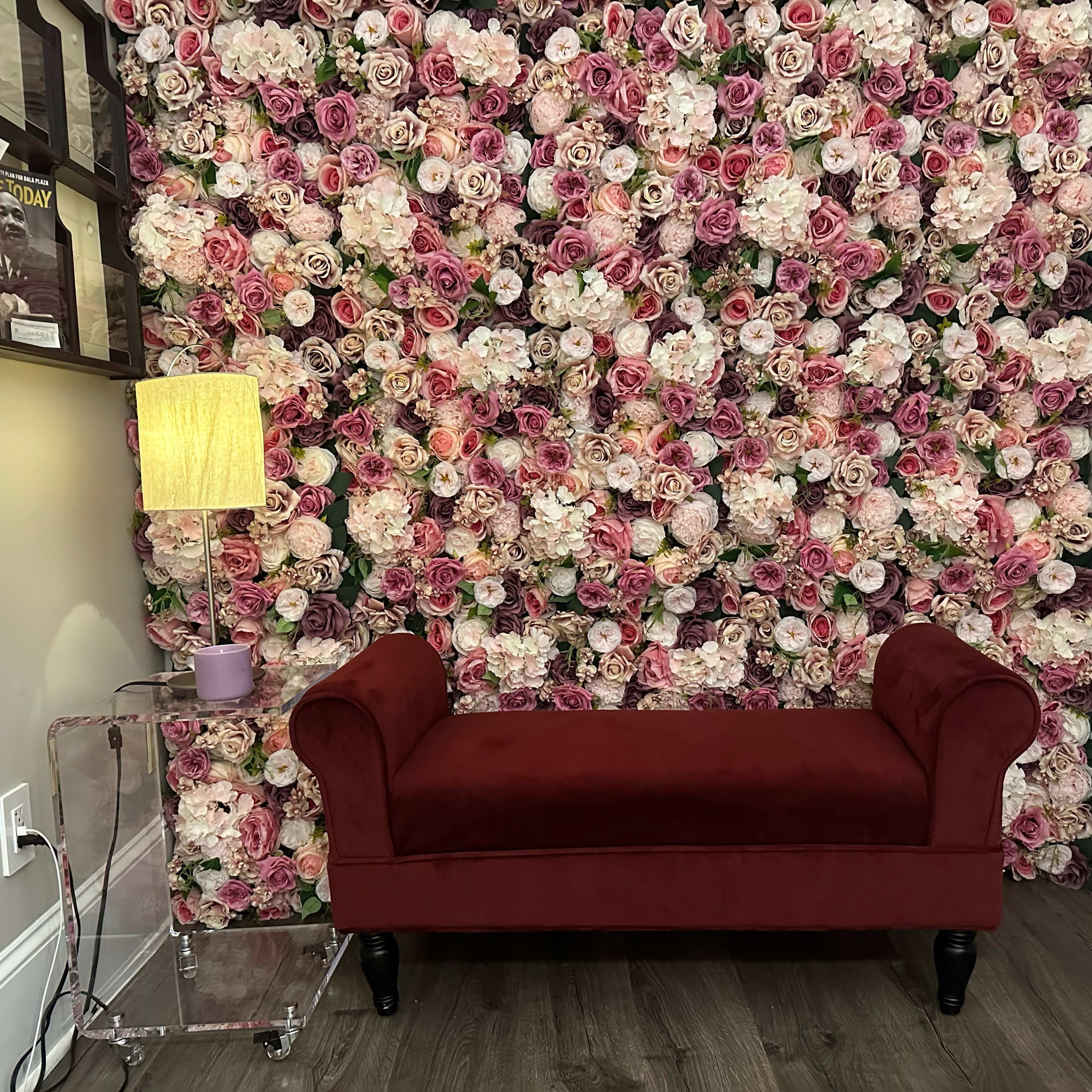 A velvet red bench in front of a wall covered with a dense arrangement of pink and white artificial flowers.