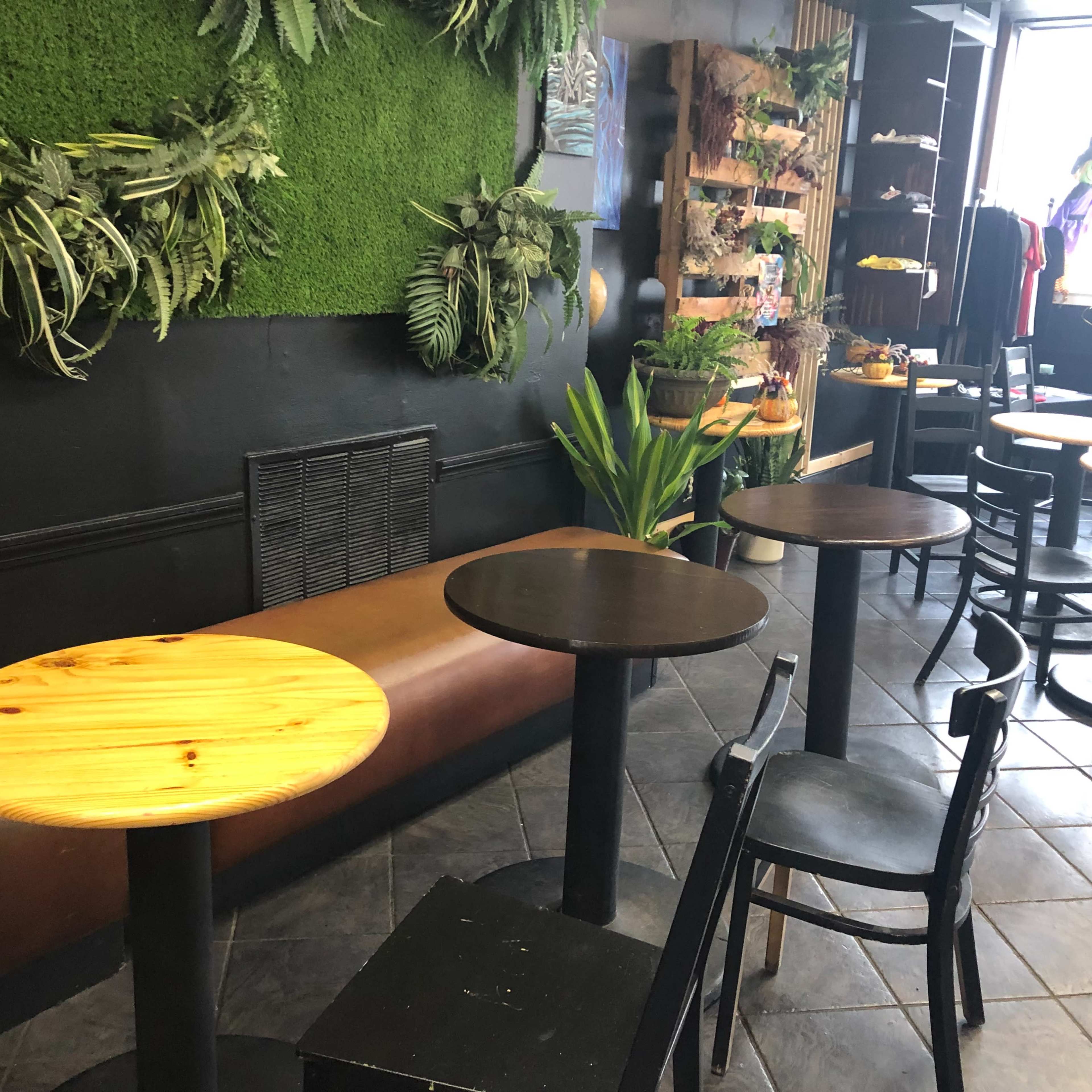 A café interior with four small round tables, a seating area on the side, and green plants on the wall.