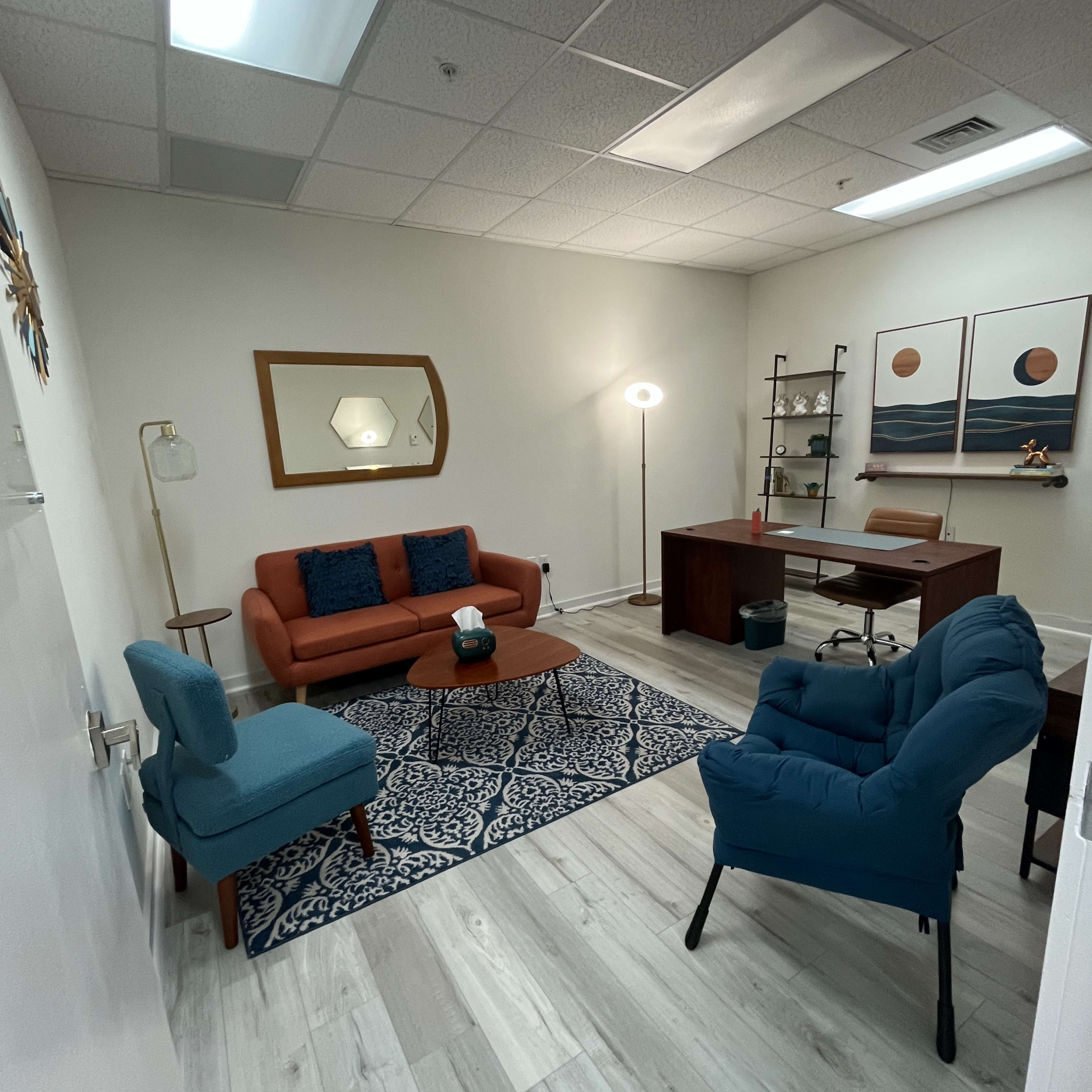 A modern office space featuring a brown desk, a blue rug, a seating area with an orange couch and two blue chairs, and wall art above the desk.