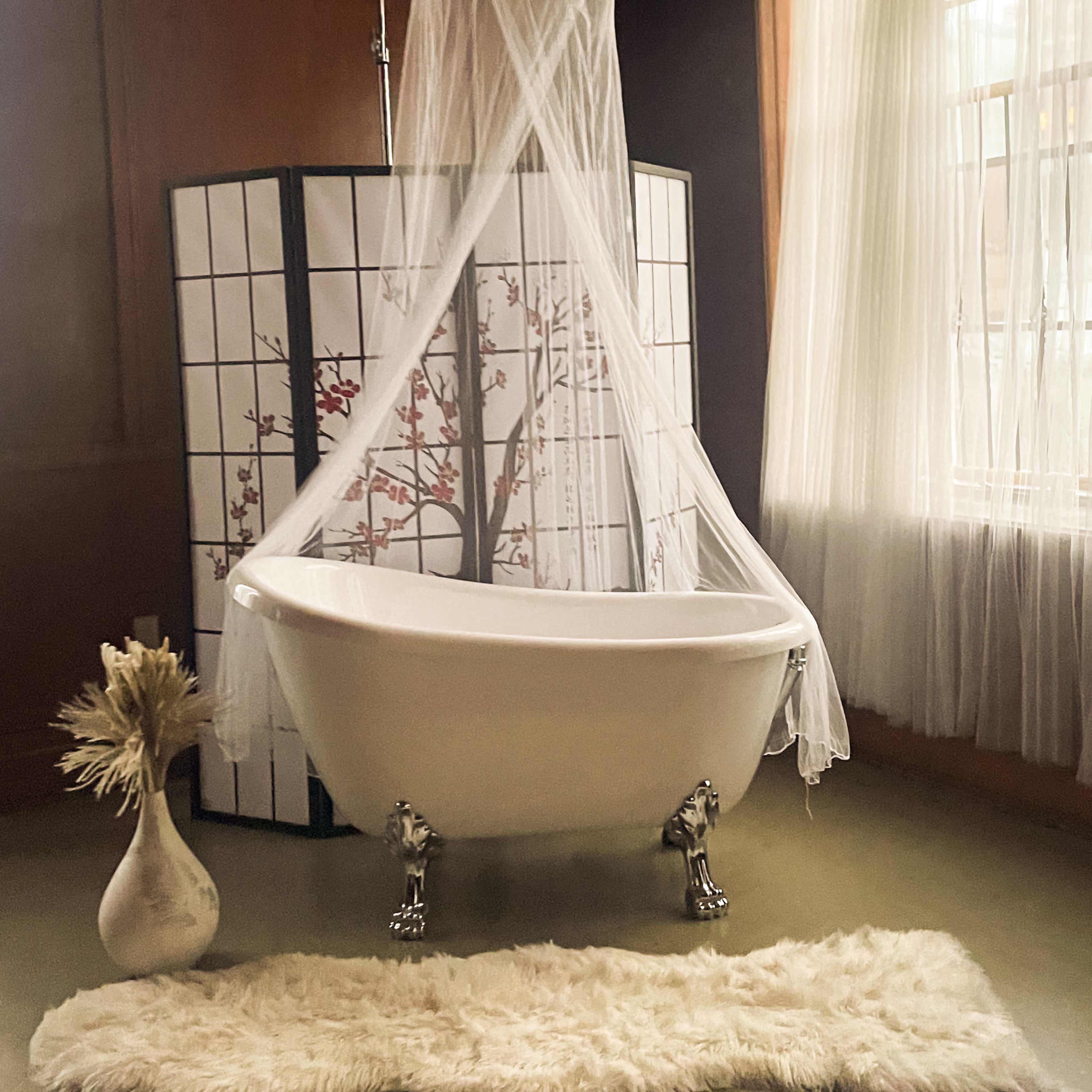 A freestanding clawfoot bathtub is positioned in a room with natural light, surrounded by a sheer canopy and a decorative screen, with a vase and a plush rug nearby.