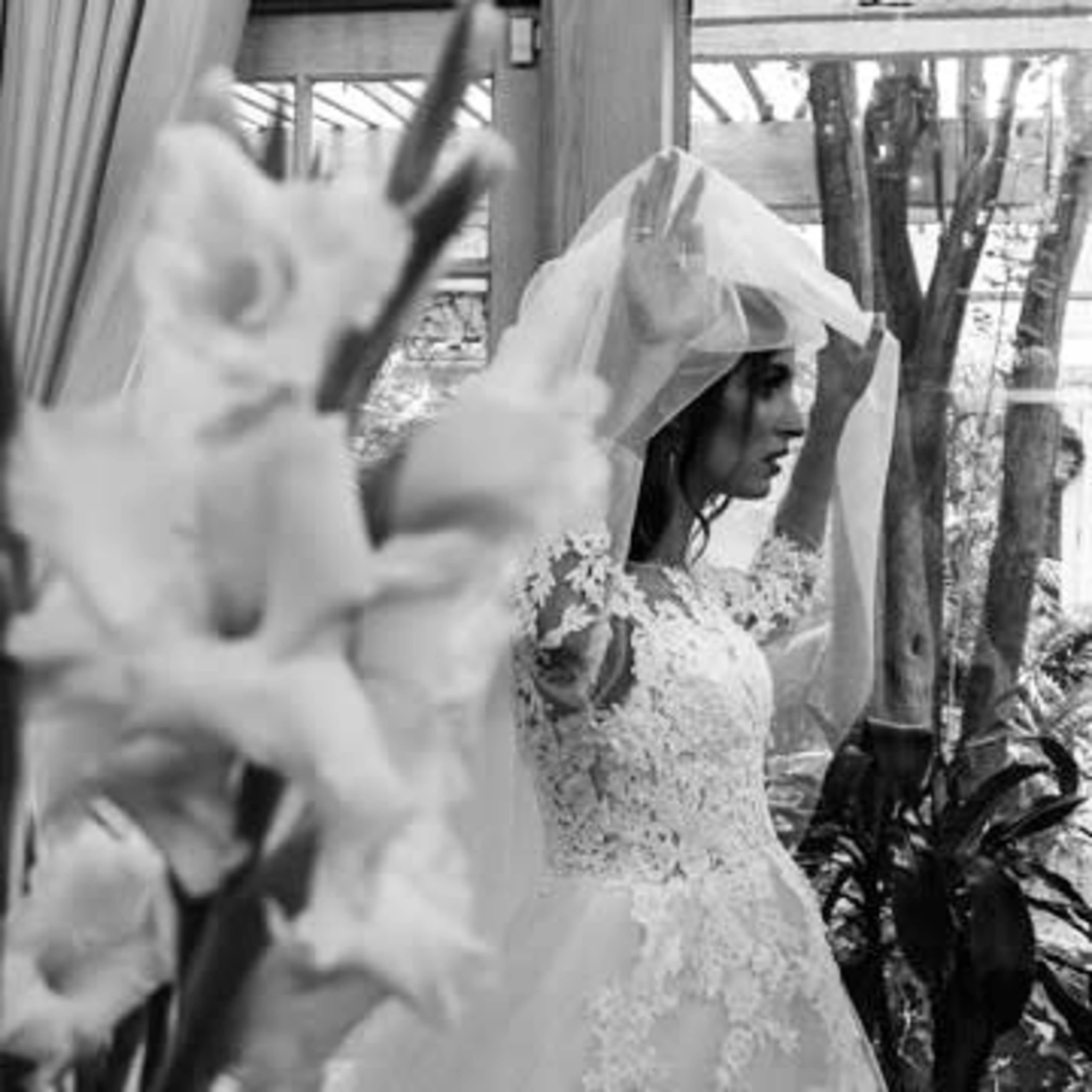A bride adjusts her veil while standing beside a floral arrangement in a glass-enclosed setting.