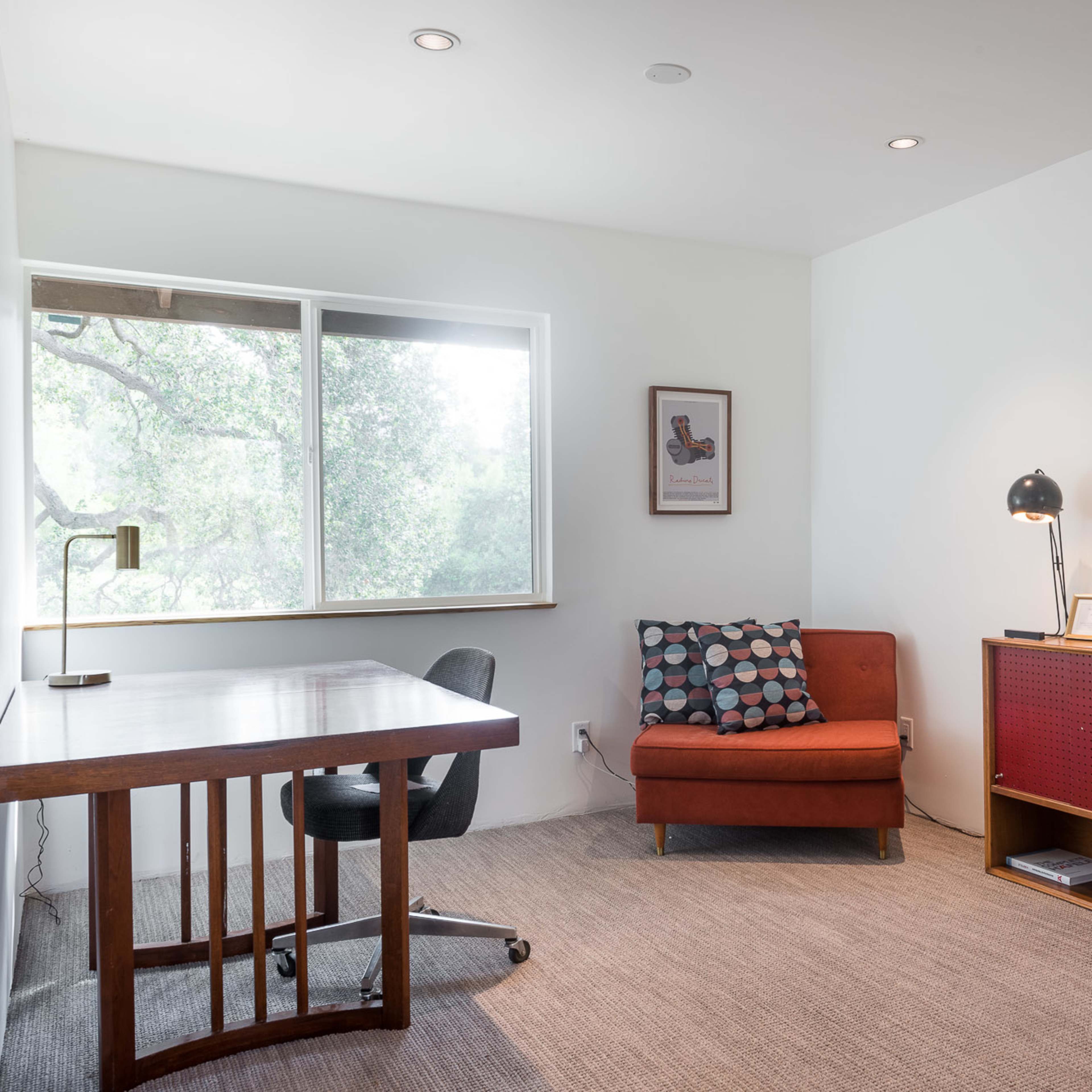 A minimalistic office space with a wooden desk, a red sofa, and a window overlooking greenery.