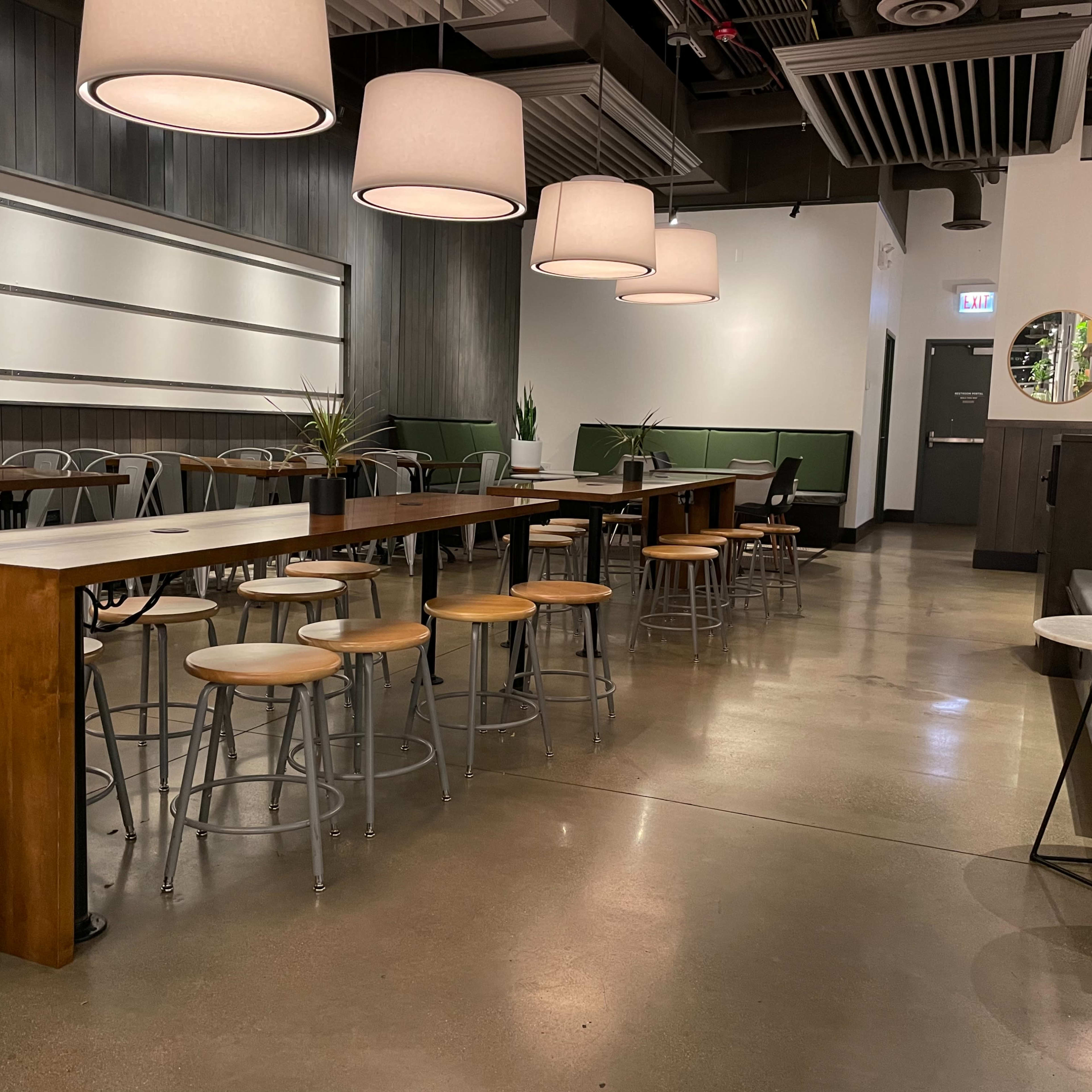 A modern cafe interior featuring wooden tables, metal stools, and large pendant lights, with plants placed around the space.