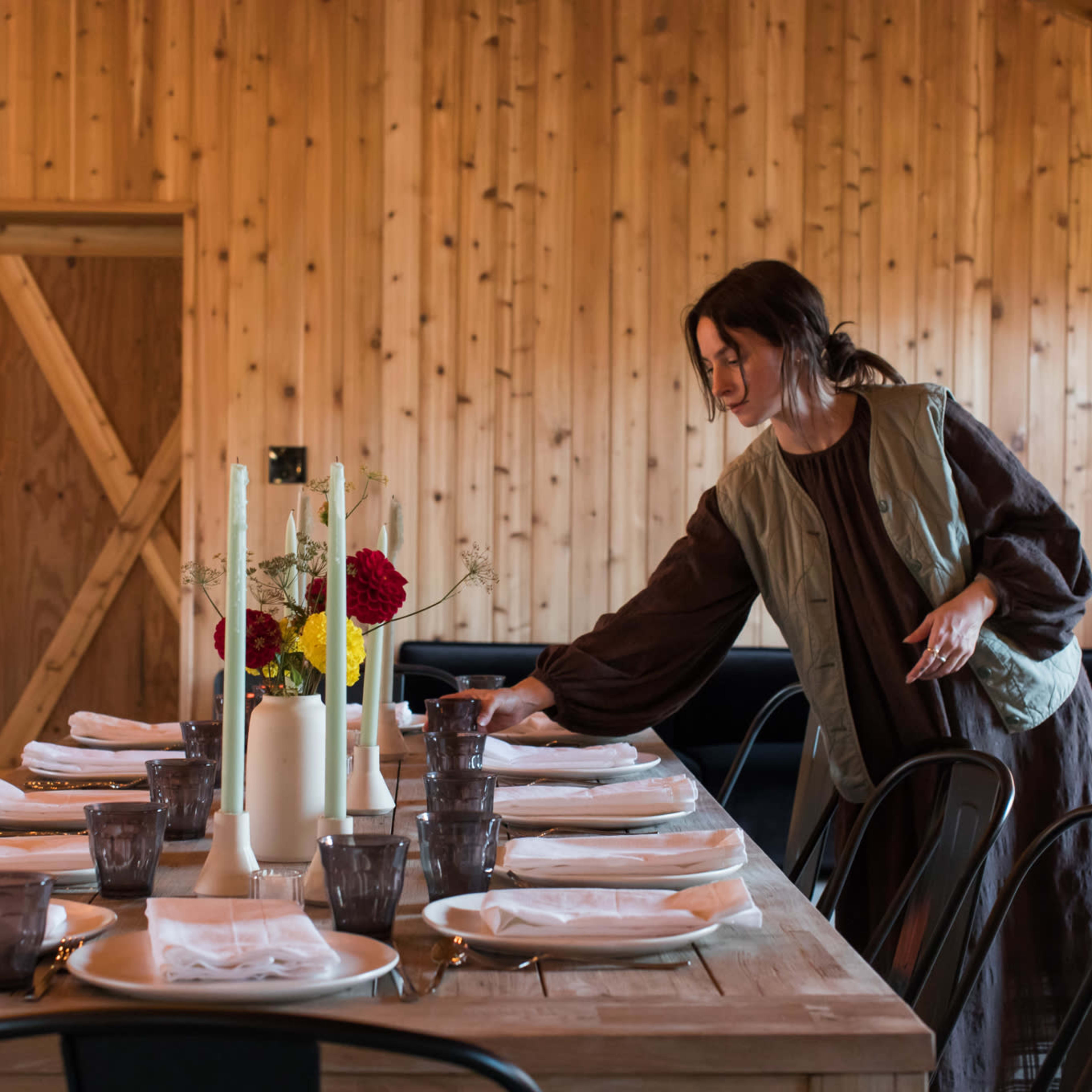 A woman adjusts the table setting in a wooden-paneled dining area with a large table and black chairs.