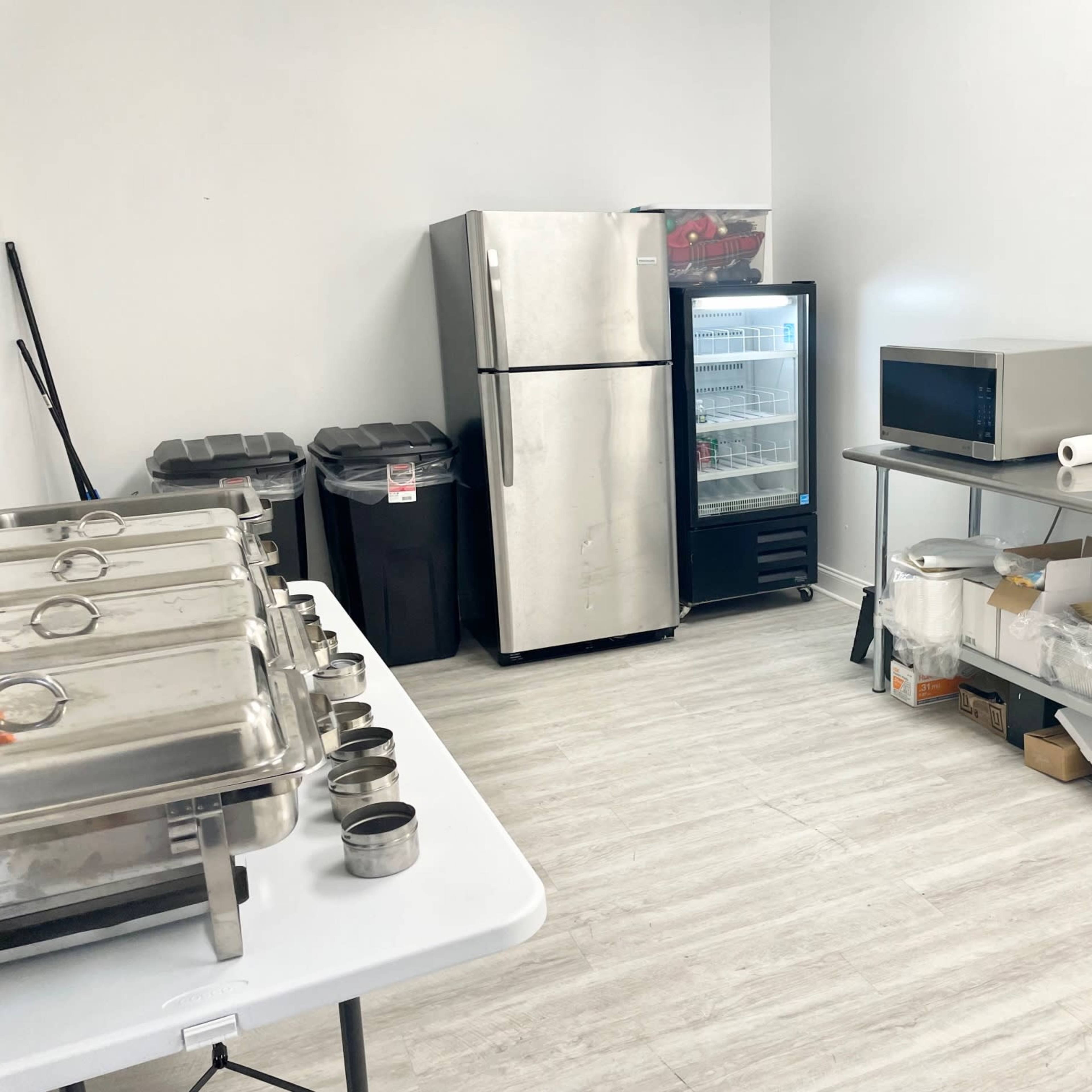 A clean kitchen space with a metal table, a microwave, a refrigerator, and several stainless steel food containers.