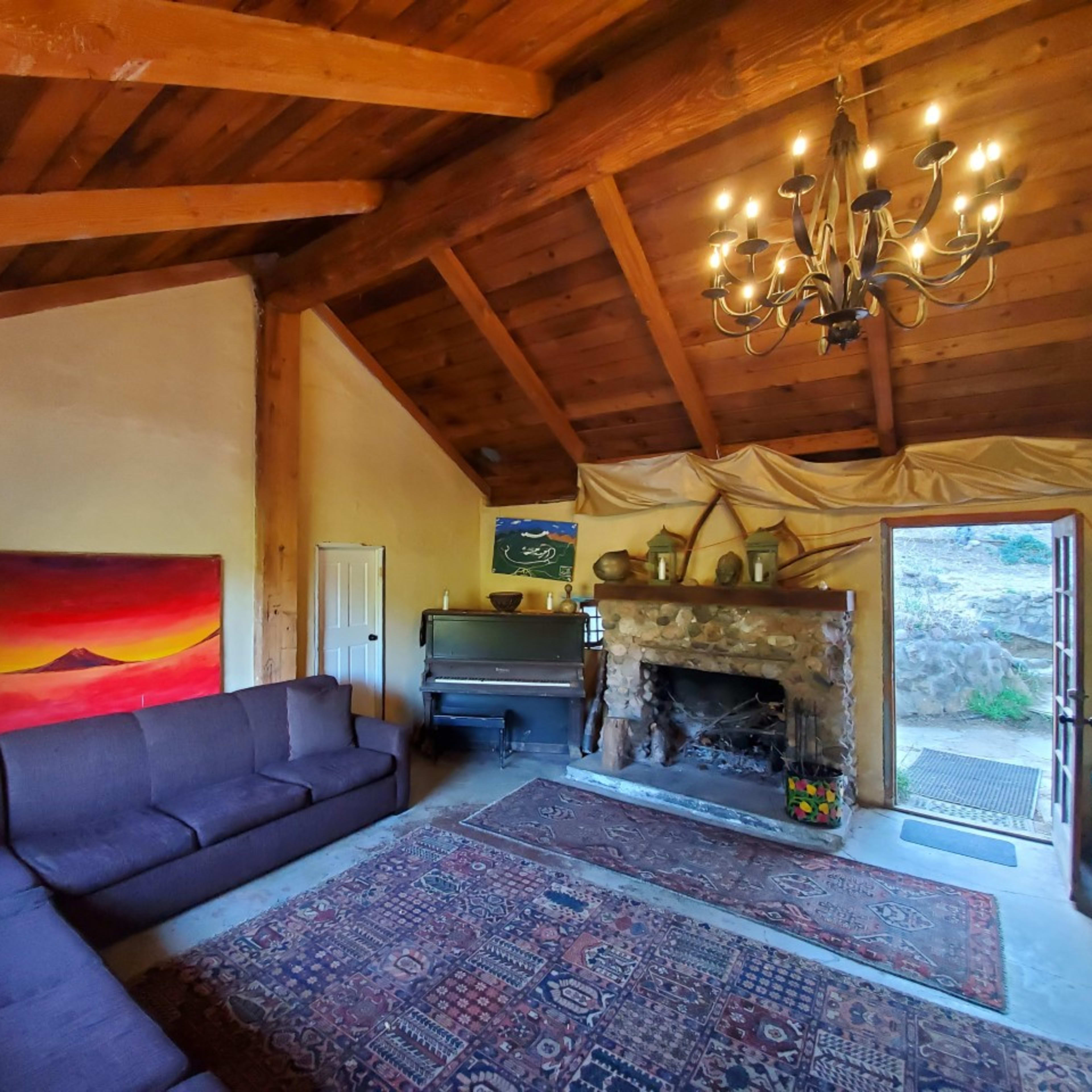 A cozy living room with a stone fireplace, a large dark sofa, and wooden beams on the ceiling, leading to an outdoor area.