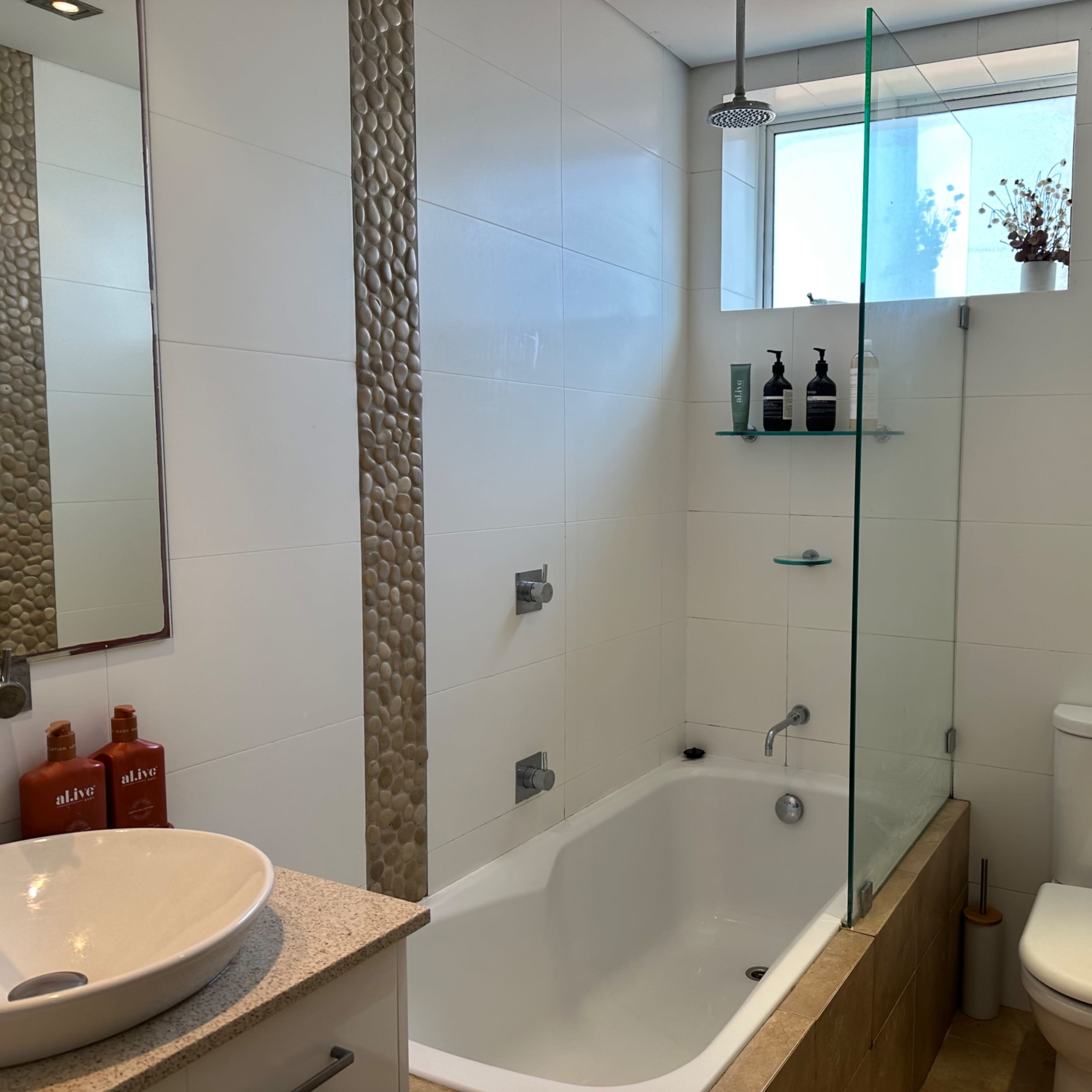 The bathroom features a white tub with a modern shower, a glass partition, and a window, complemented by a countertop sink and a toilet.