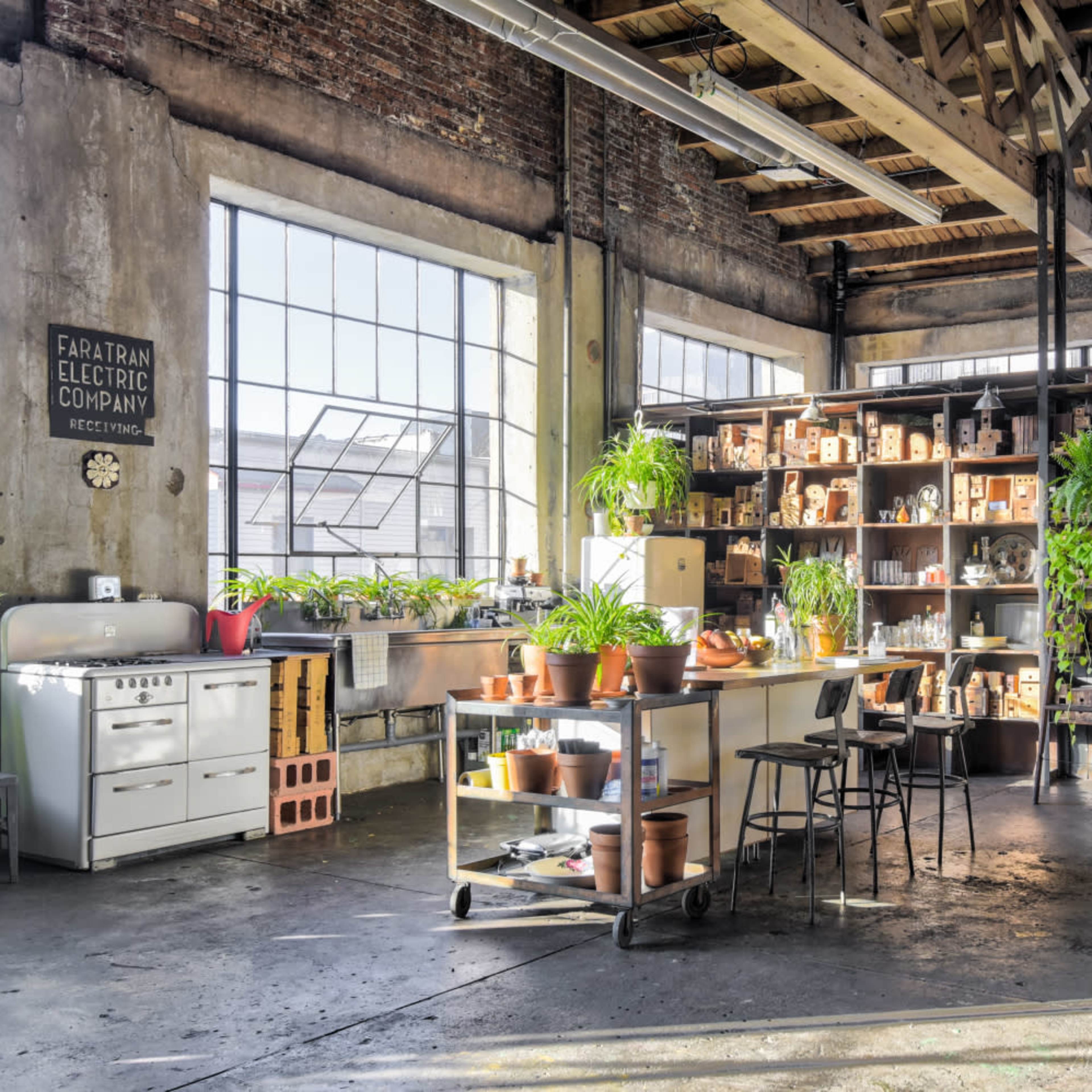 An industrial-style interior featuring a spacious workspace with shelves filled with plants and various containers, along with a kitchen area and large windows.