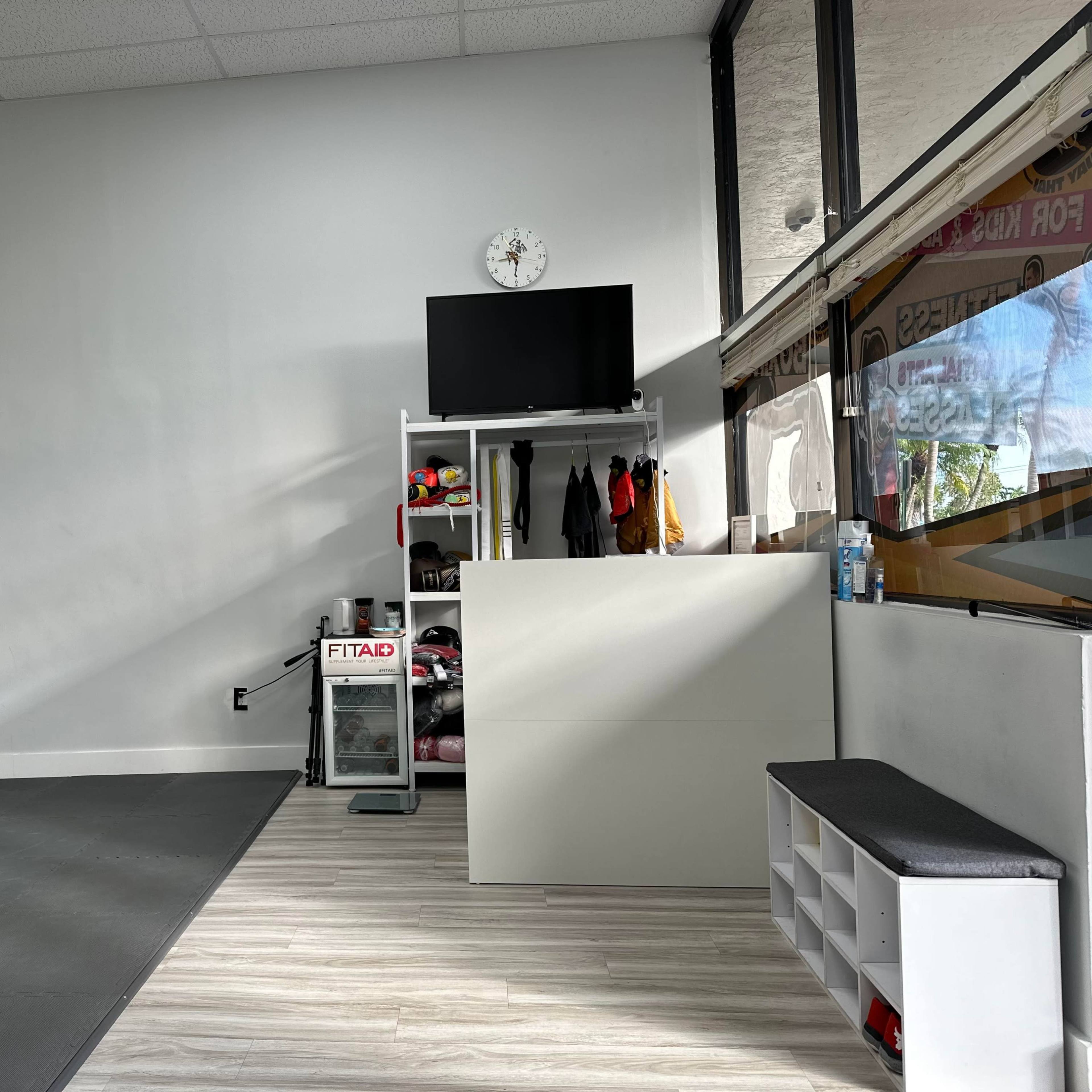 A well-lit, minimalist interior of a fitness studio featuring a television, storage shelves with equipment, a clock on the wall, and a black mat on the floor.