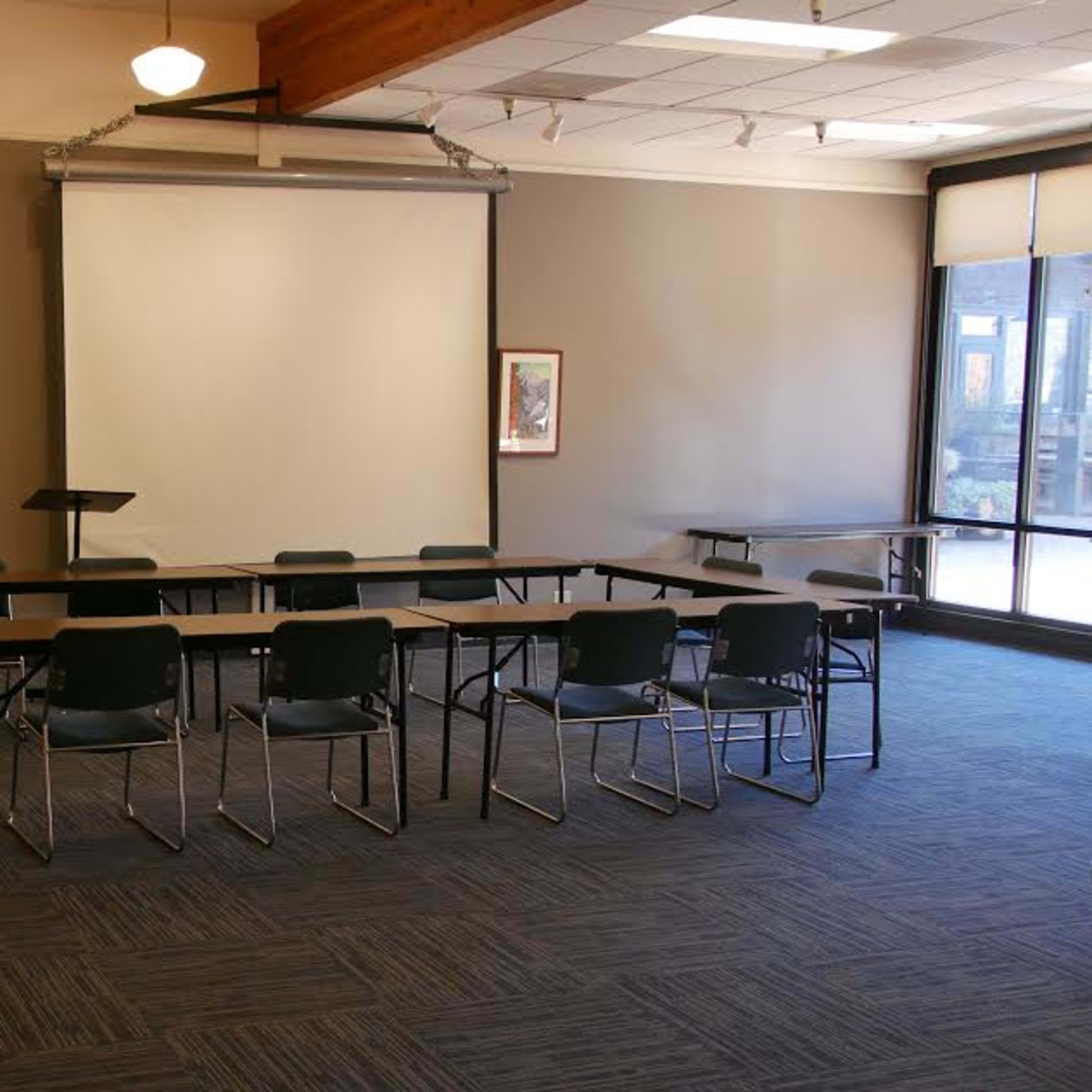 A meeting room features a central arrangement of tables and chairs, with a projector screen and framed artwork on the walls.