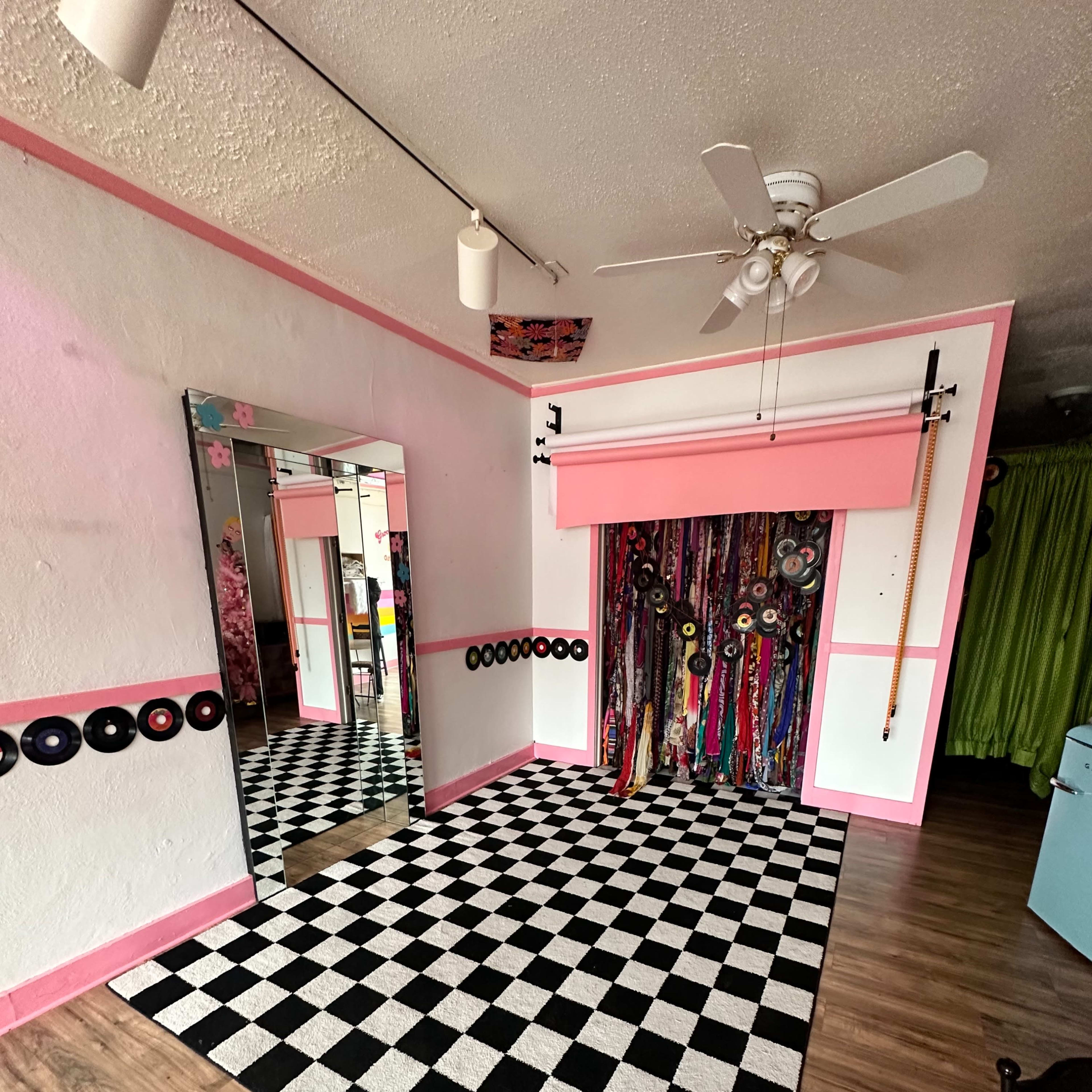 The room features a checkered black-and-white floor, a large mirror, and a colorful wall of fabric drapes, with pink accents throughout.