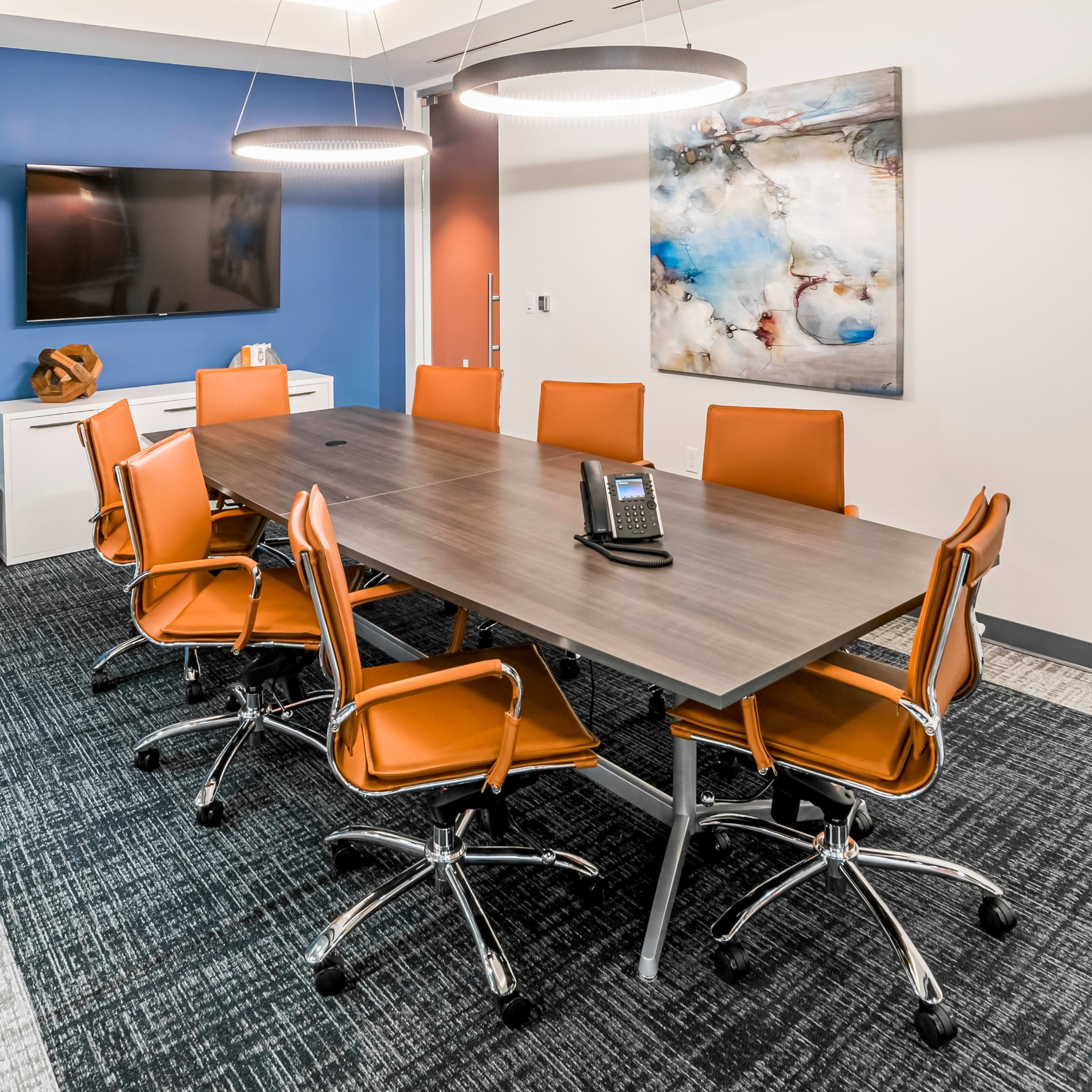 A modern conference room features a long wooden table surrounded by orange chairs, with dimmable pendant lights above and a TV mounted on the wall.