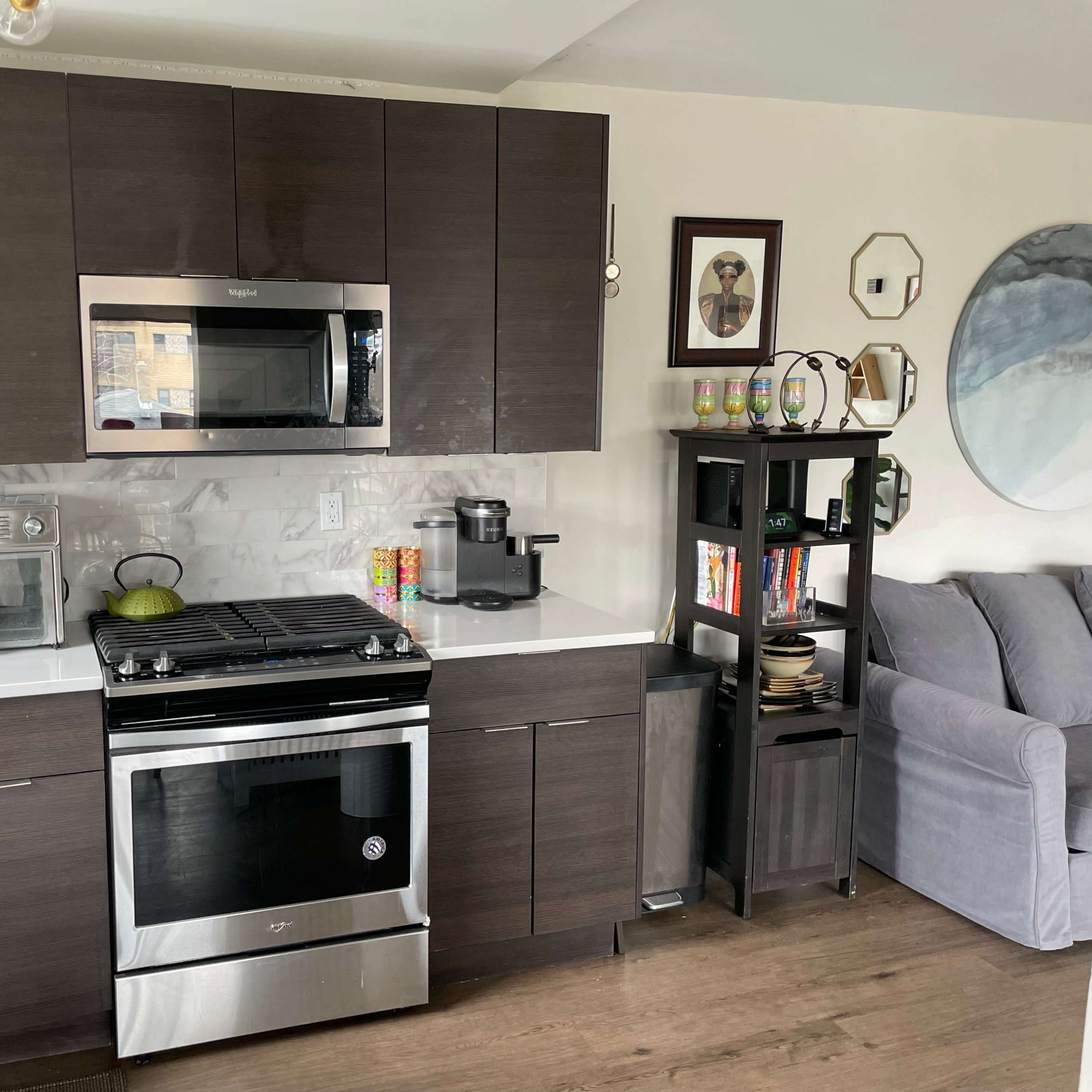 A modern kitchen with dark cabinetry, a stainless steel gas stove, a microwave, a coffee maker, and a small bookshelf beside a gray couch.
