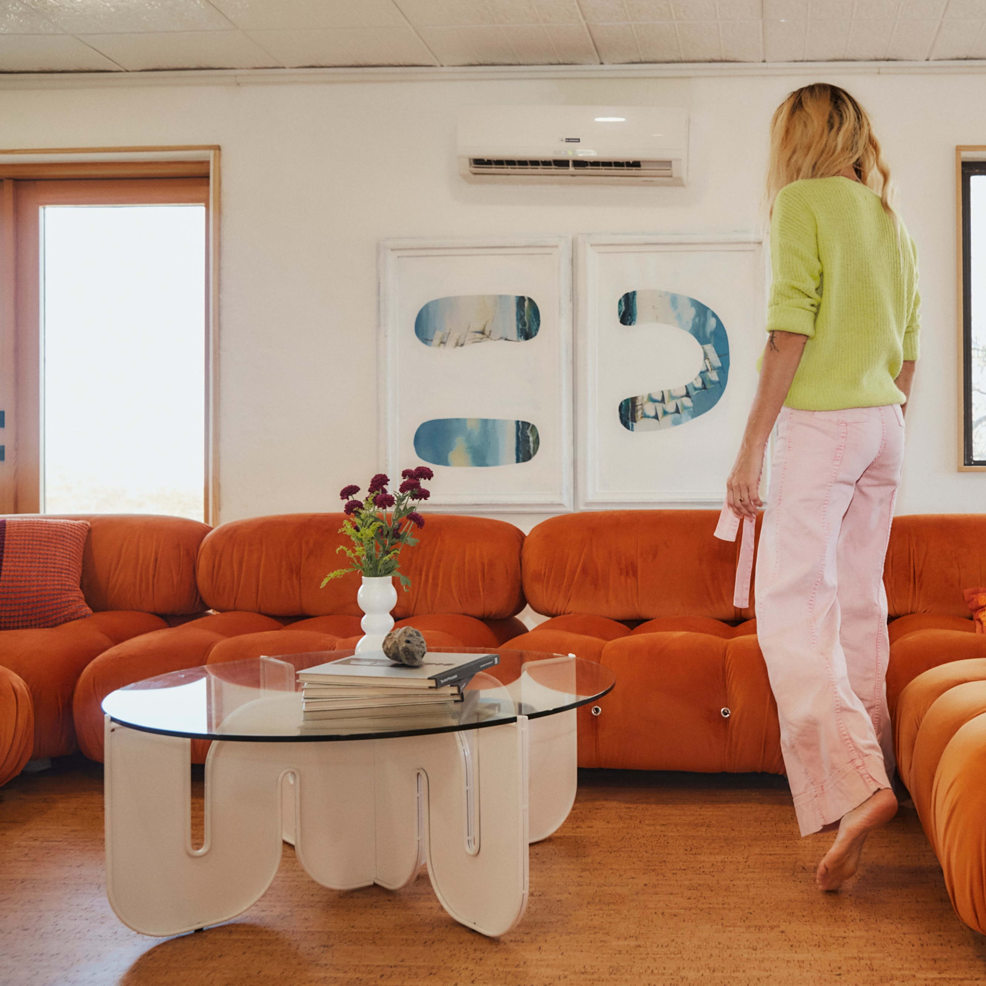 A person walks barefoot on a beige floor beside an orange couch and a glass coffee table in a well-lit room with large windows.