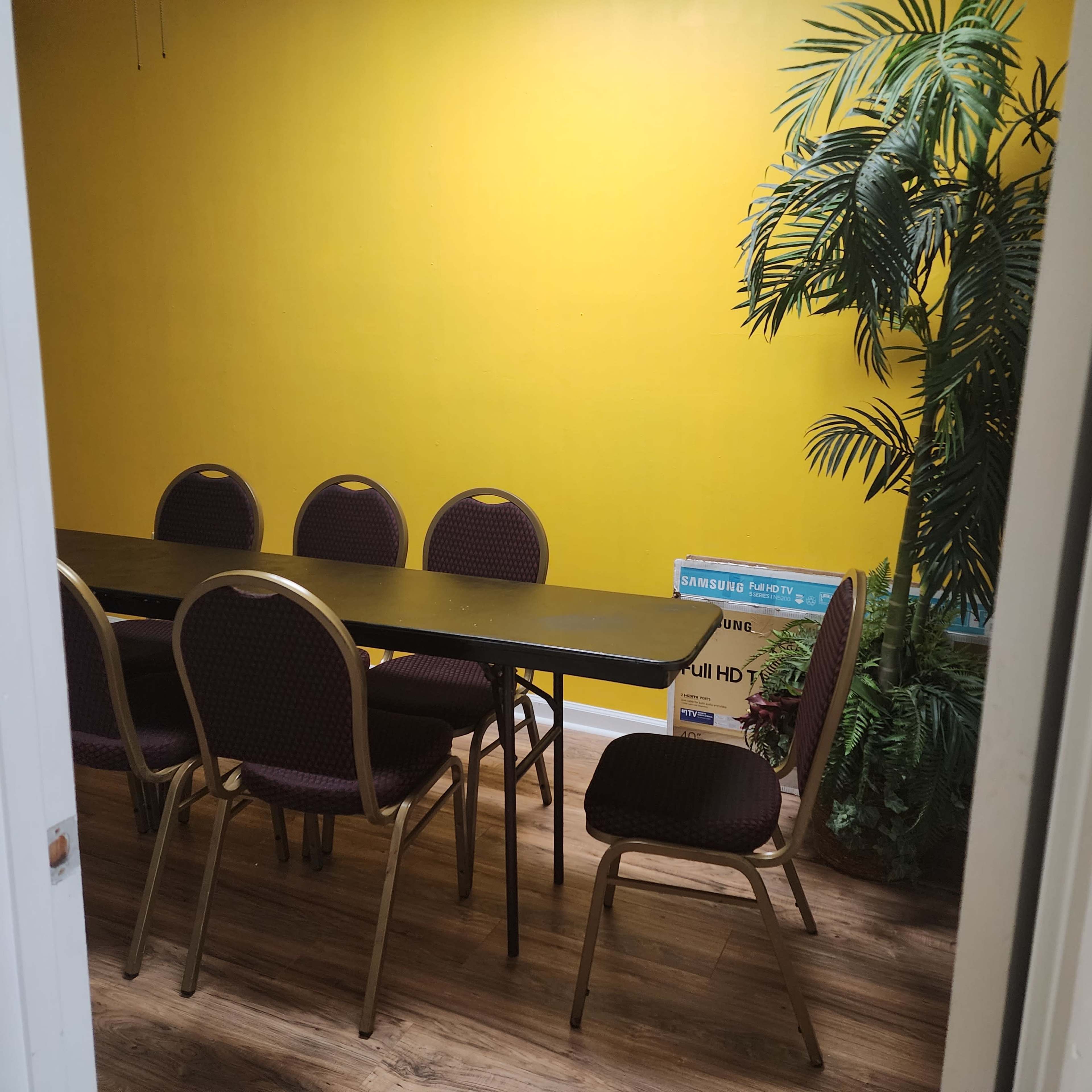 A room with a yellow wall features a long black table surrounded by six purple chairs and a green potted plant.
