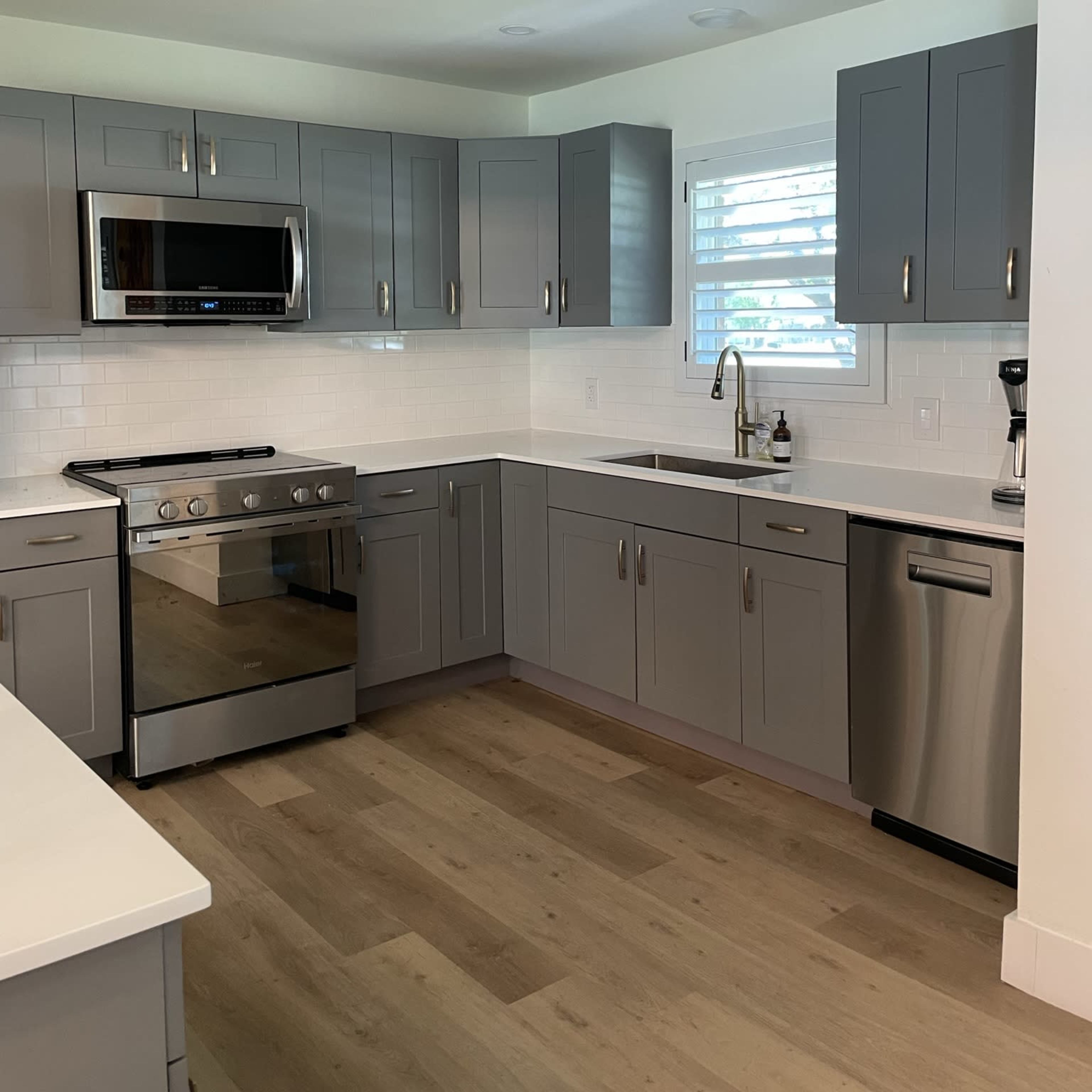 A modern kitchen featuring gray cabinets, stainless steel appliances, and a white countertop with a sink and window.