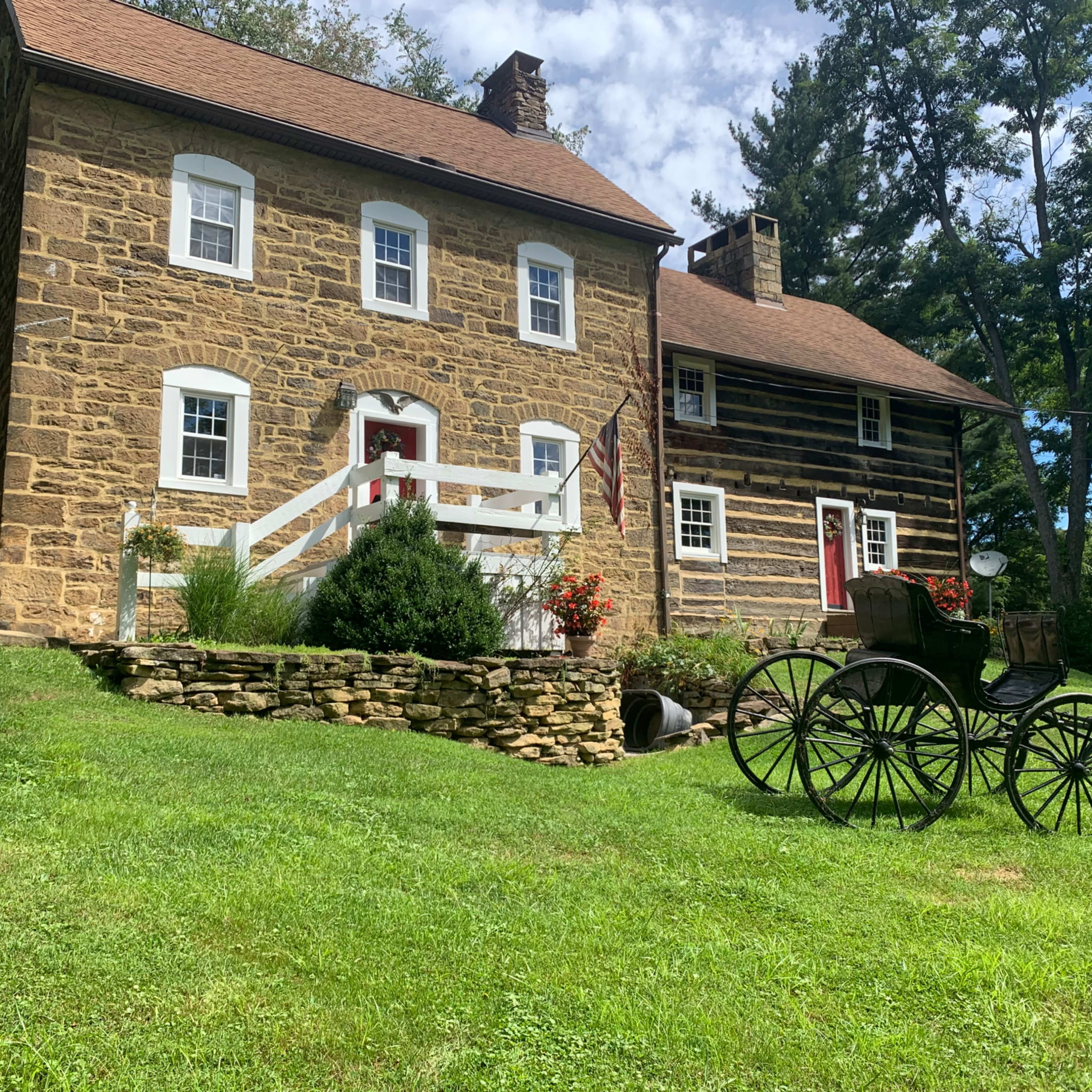 Two historic homes, one made of stone and the other of wood, with a grassy area and a vintage horse-drawn carriage in the foreground.