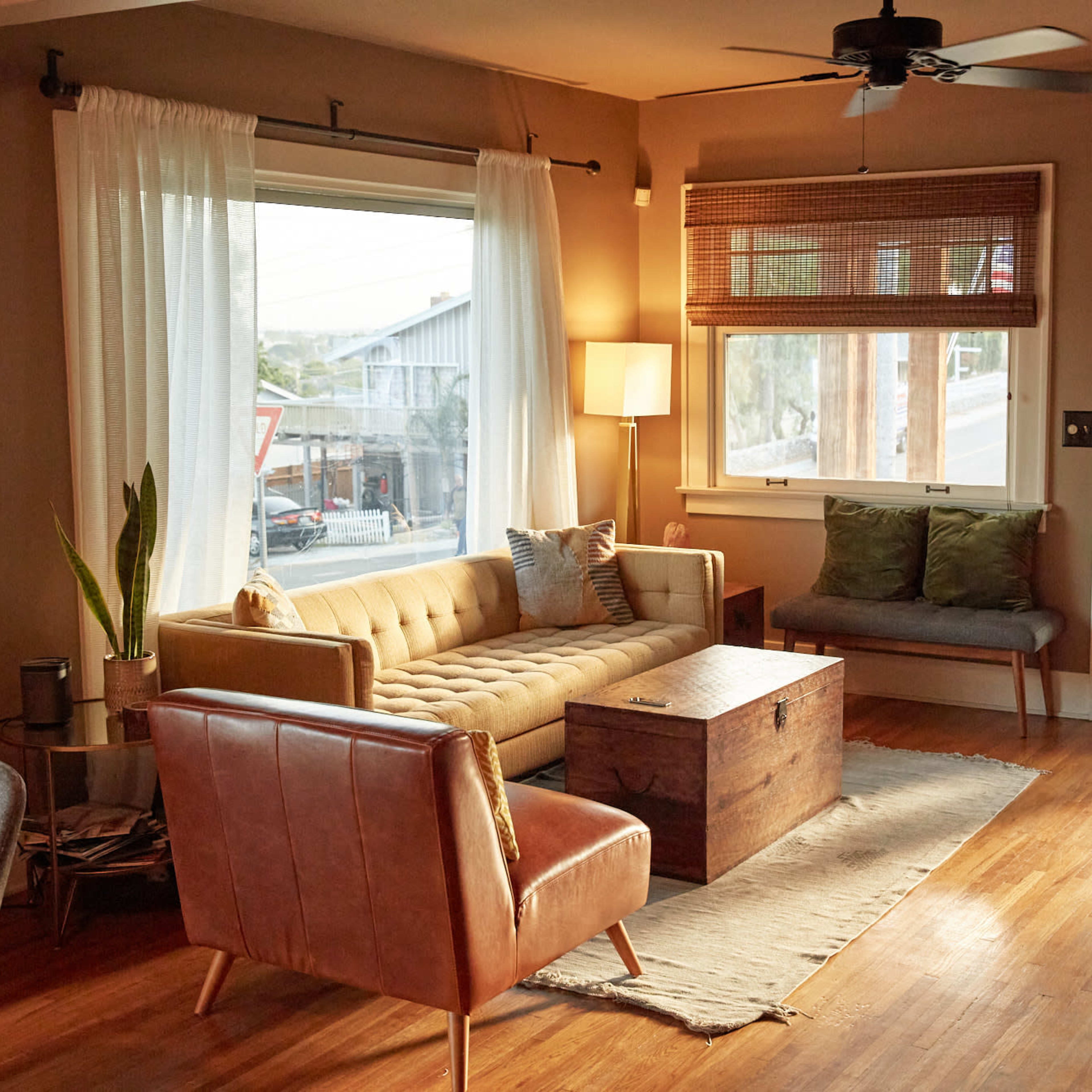 The living room features a tan tufted sofa, a wooden coffee table, and a large window with light curtains, providing a view of the outside.