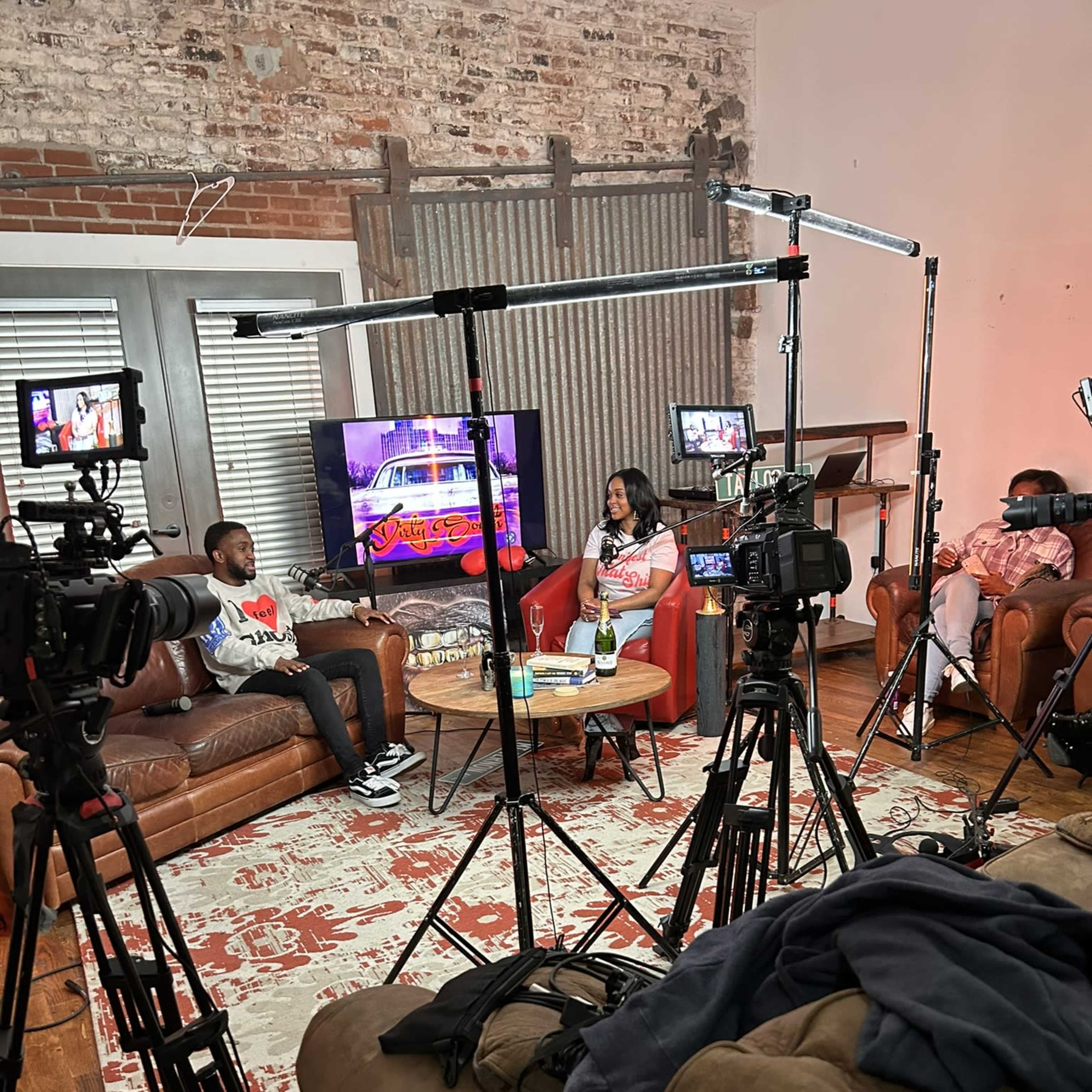 A television studio setup with cameras and lights surrounding two people seated on a couch, discussing on a set featuring a colorful backdrop.