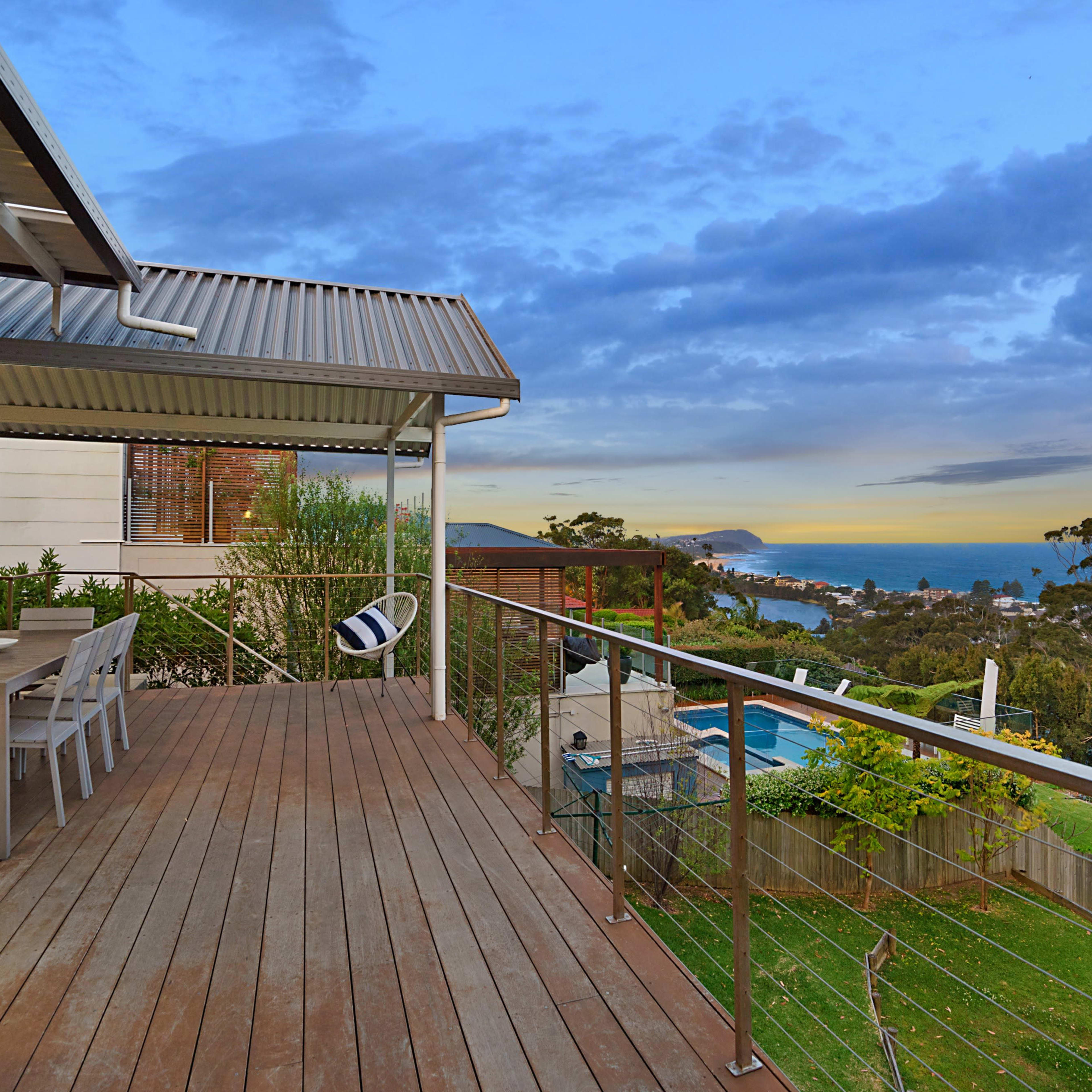 A wooden deck with a dining table overlooks a coastal view at sunset, featuring a swimming pool and surrounding greenery.