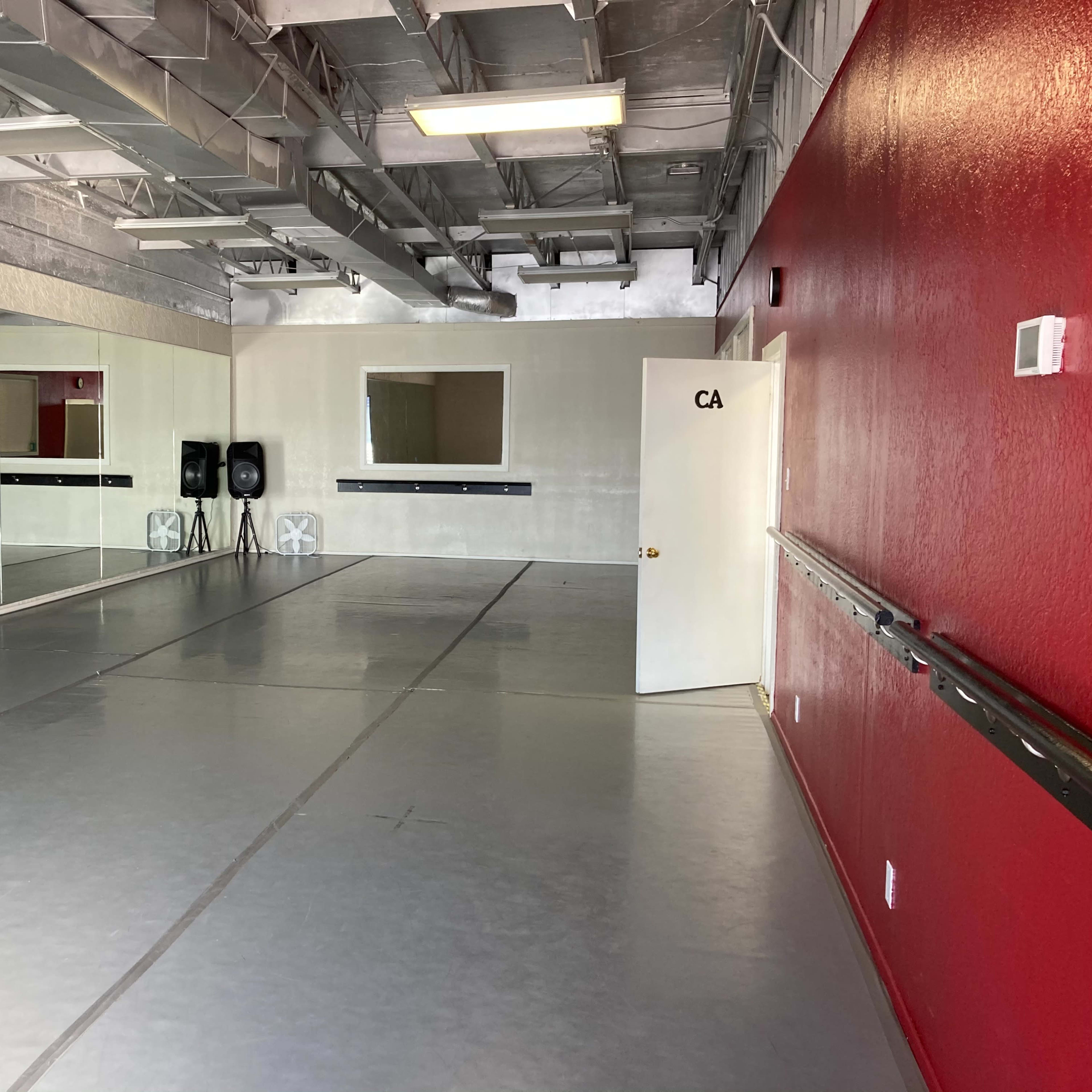 A dance studio with mirrored walls, a grey floor, and a red accent wall, featuring a door labeled "CA."