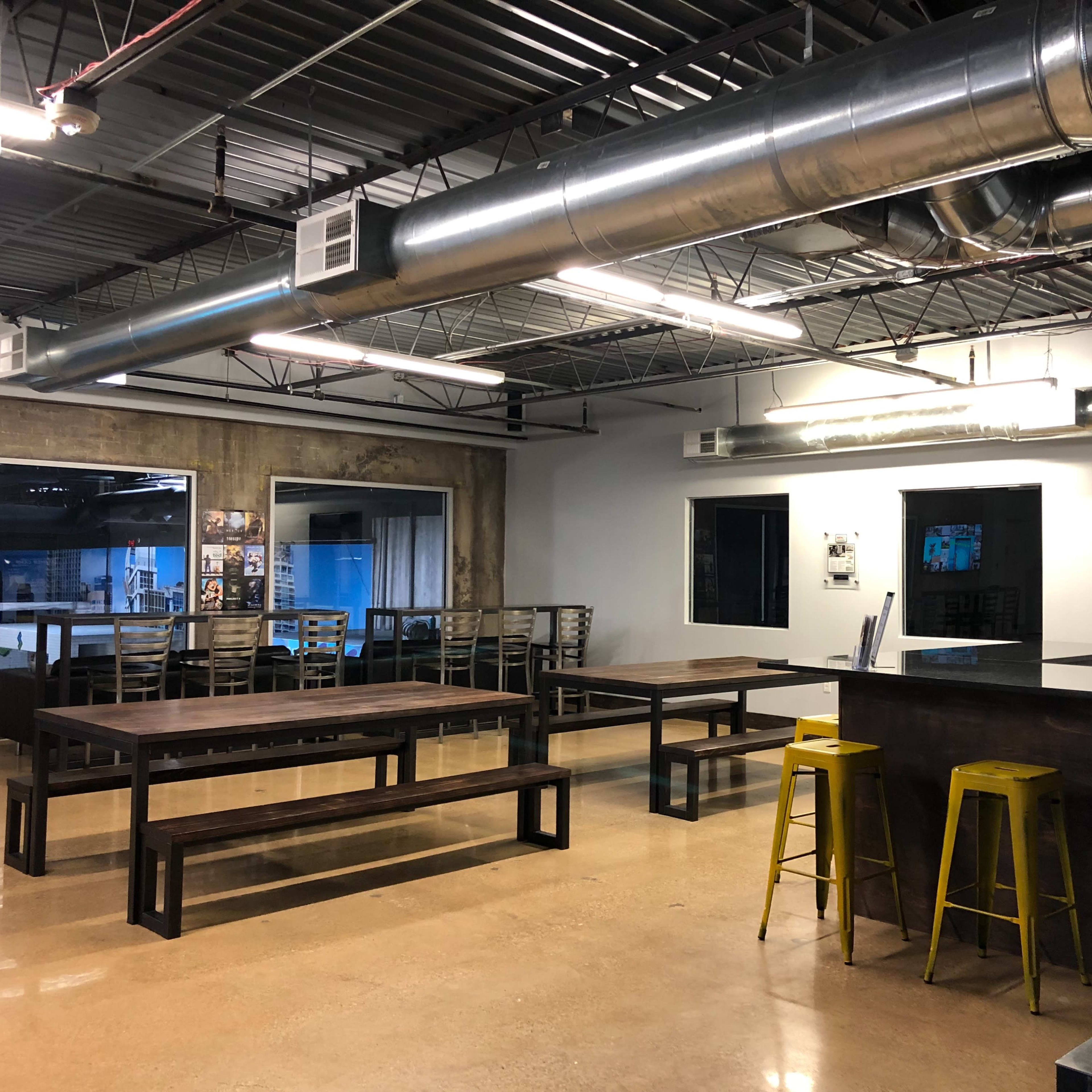 A spacious industrial-style workspace with wooden tables, metal bar stools, and exposed ductwork on the ceiling.