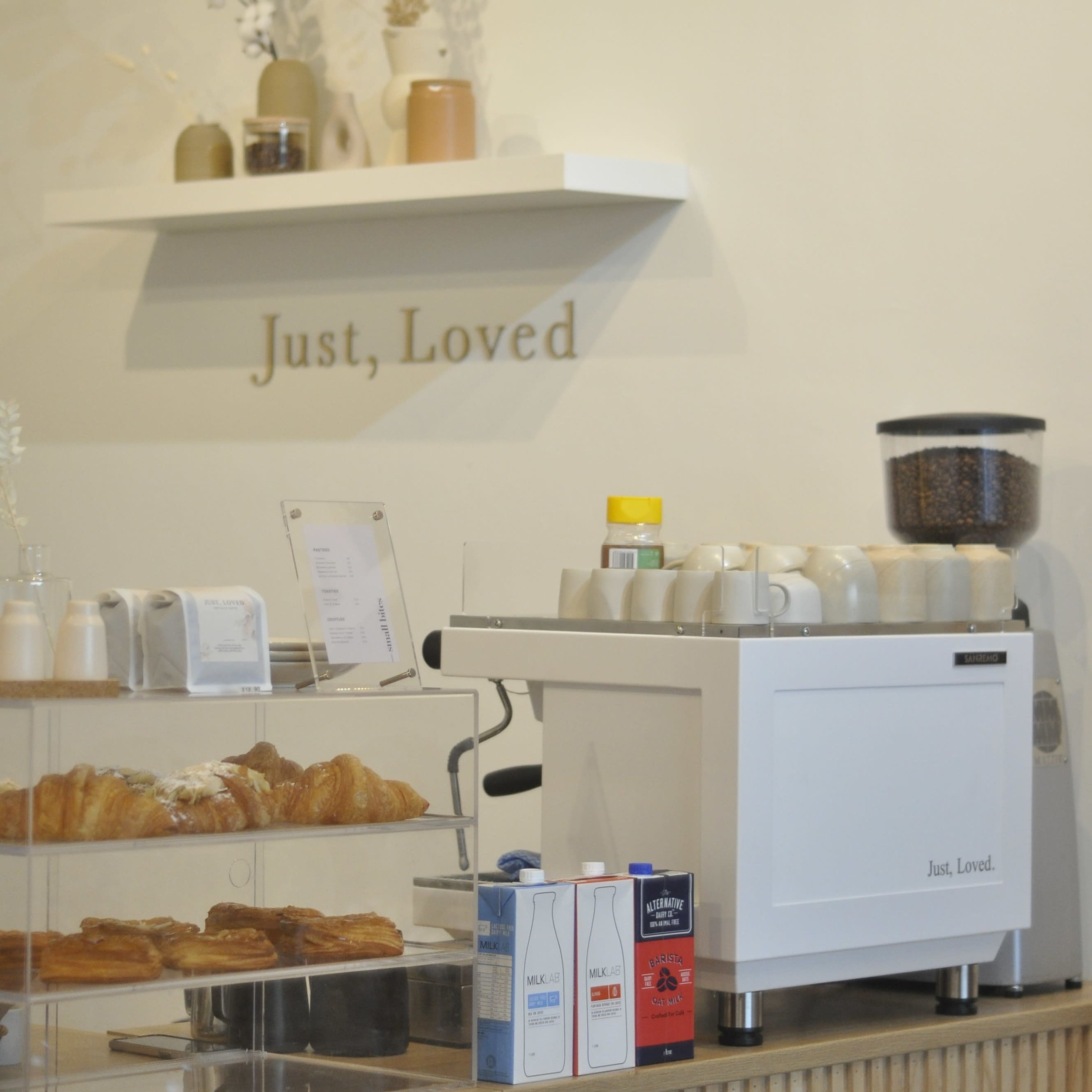 A coffee shop counter features a display of pastries, a coffee machine, and decorative elements on the wall.