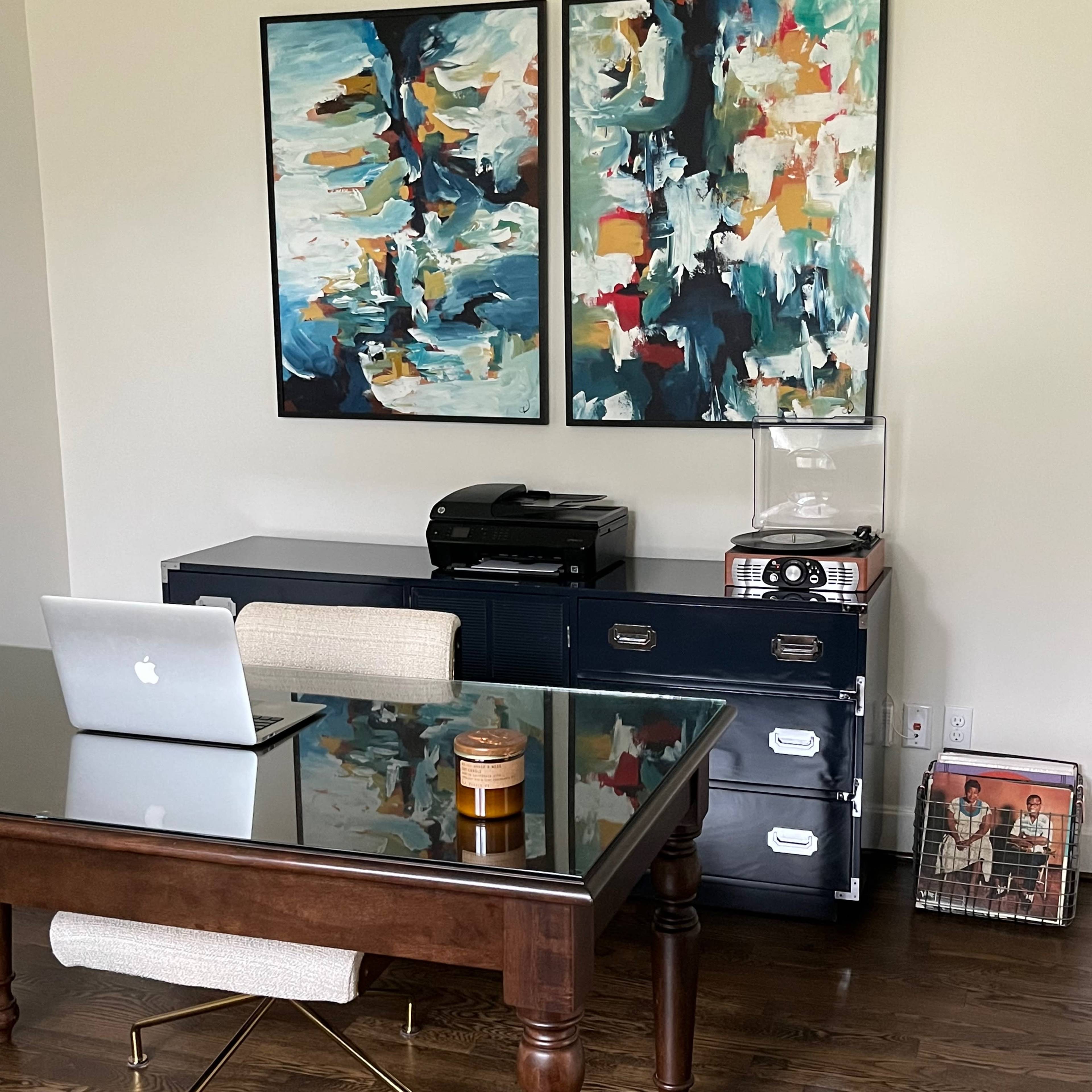 A modern office space features a wooden desk with a laptop, a printer and record player on a side cabinet, and two colorful abstract paintings on the wall.