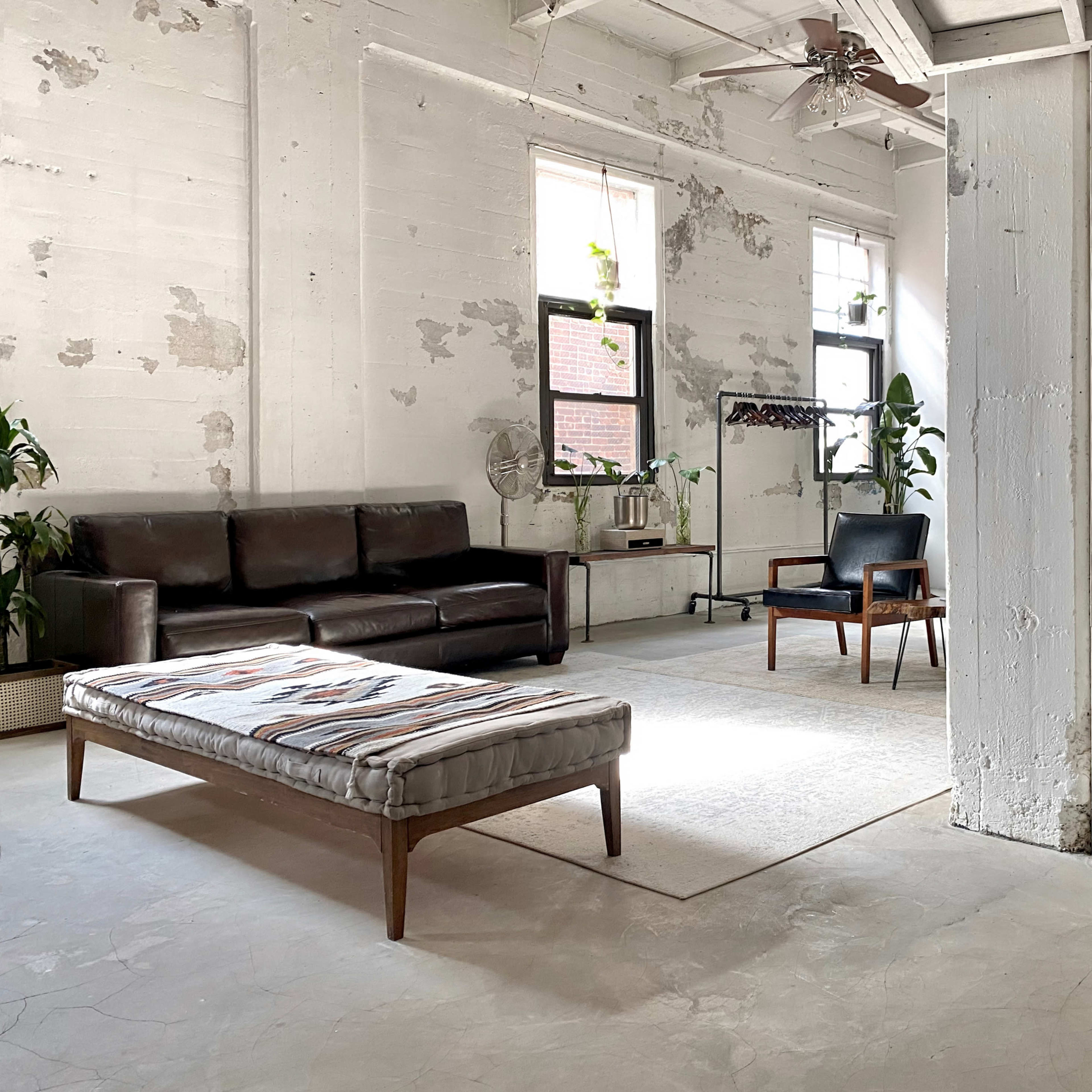 A spacious, minimalist living area with a dark sofa, two chairs, and a bed-style bench on a light floor, surrounded by large windows.