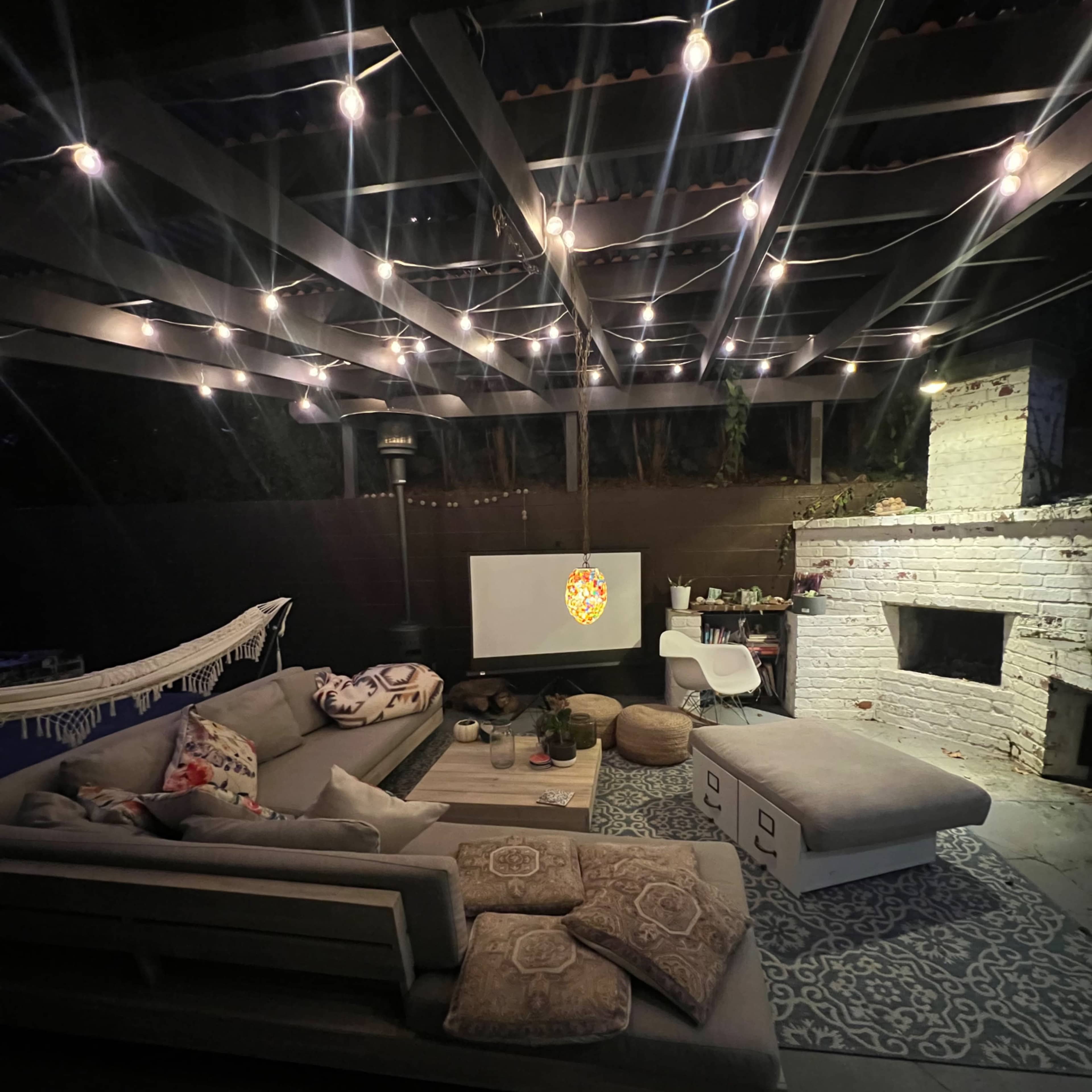 A cozy outdoor living area features a sectional sofa, a large TV screen, and string lights overhead, set against a backdrop of a white brick wall.
