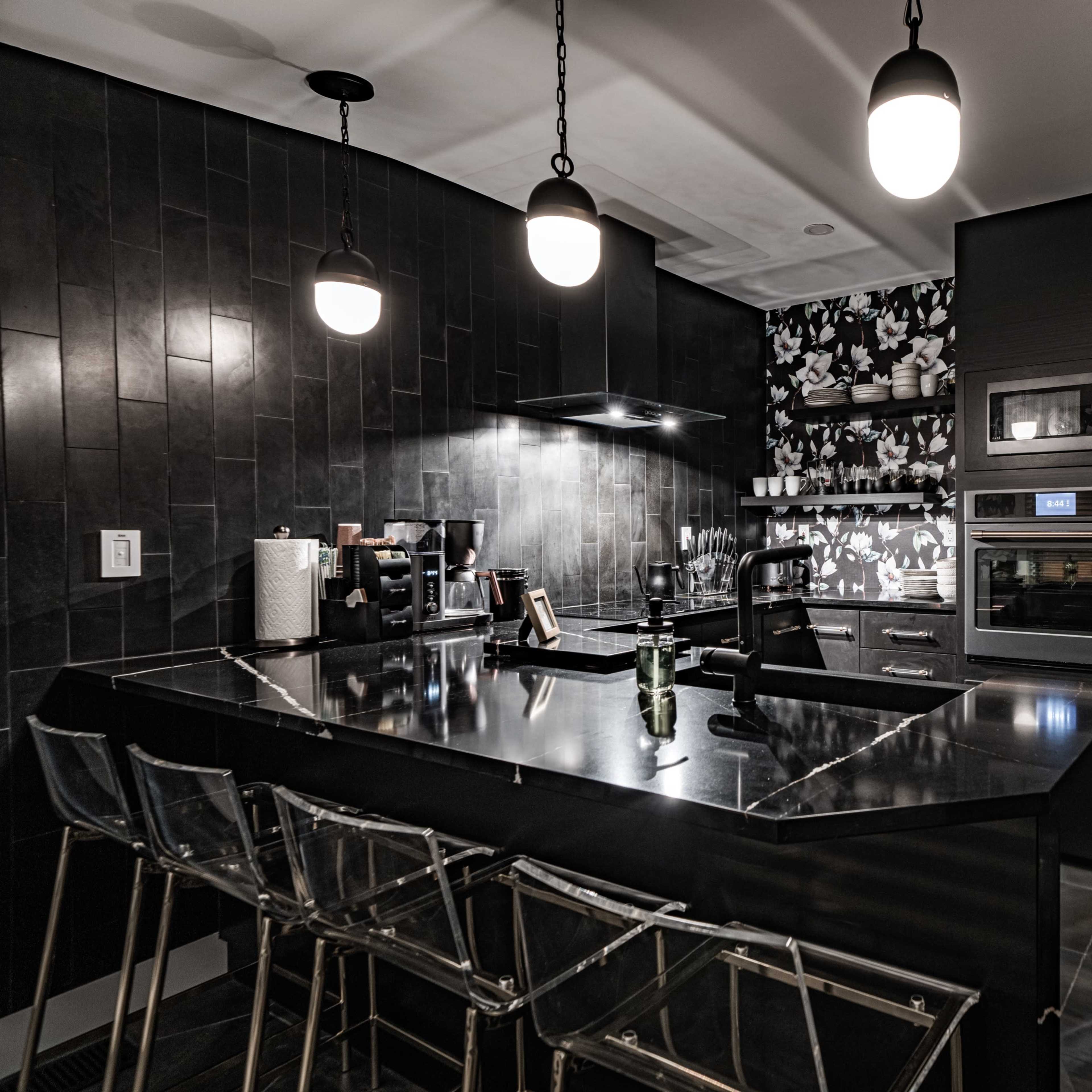A modern kitchen features dark cabinetry, sleek appliances, and a black island with transparent bar stools under pendant lighting.