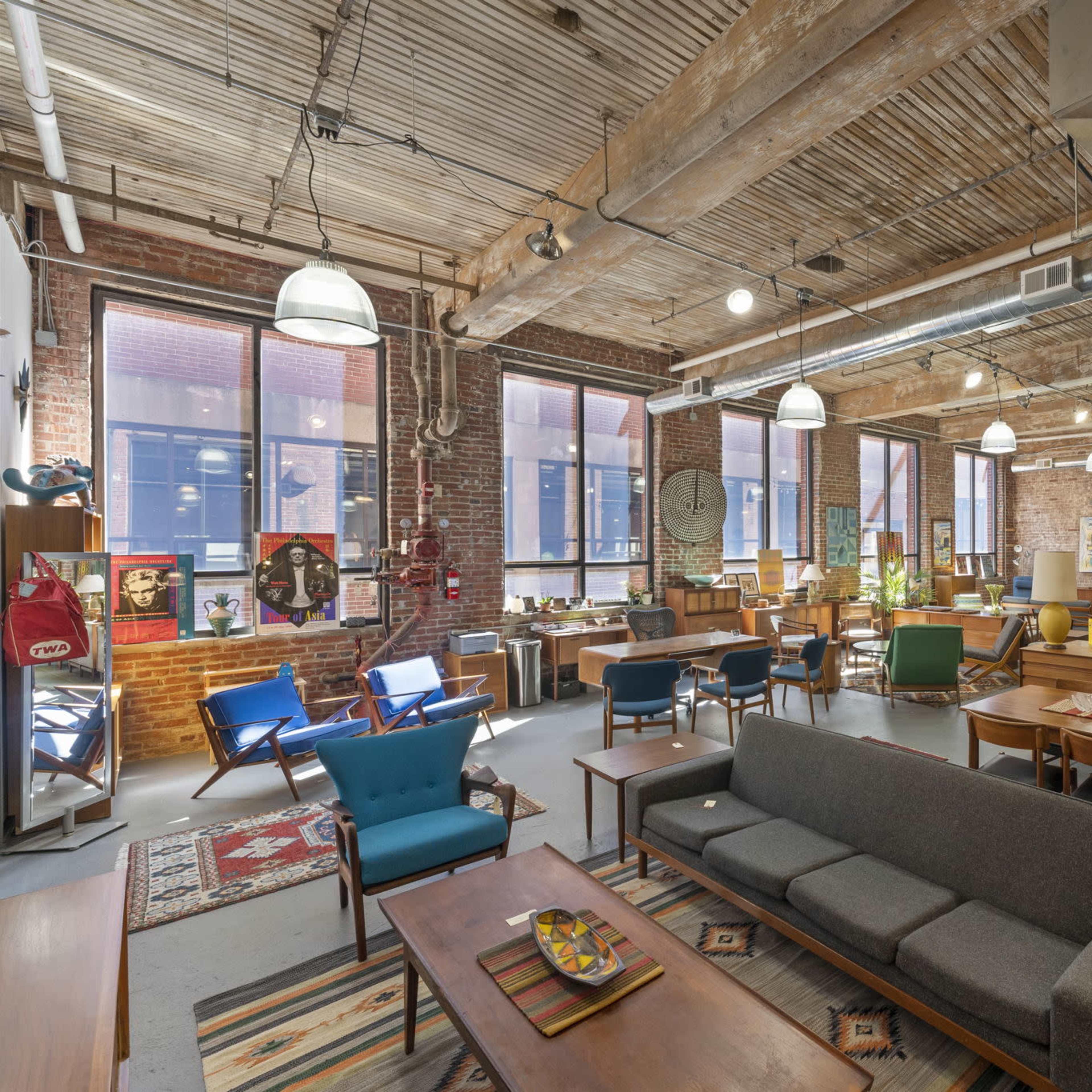 A spacious, well-lit room filled with various vintage furniture pieces, including sofas, chairs, and tables, against a backdrop of exposed brick walls and large windows.
