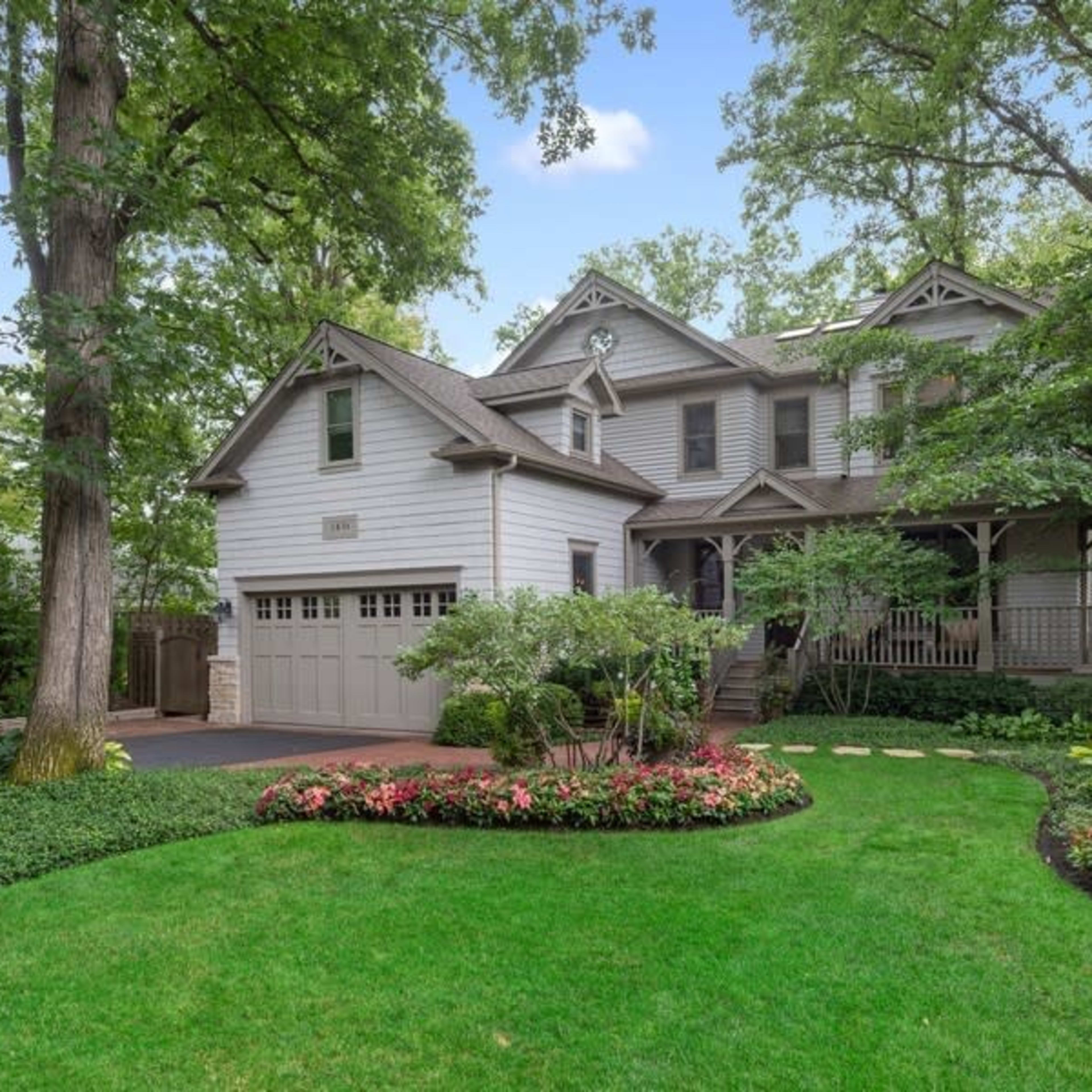 A large two-story house with a gray exterior and a covered porch is surrounded by well-manicured landscaping and a green lawn.