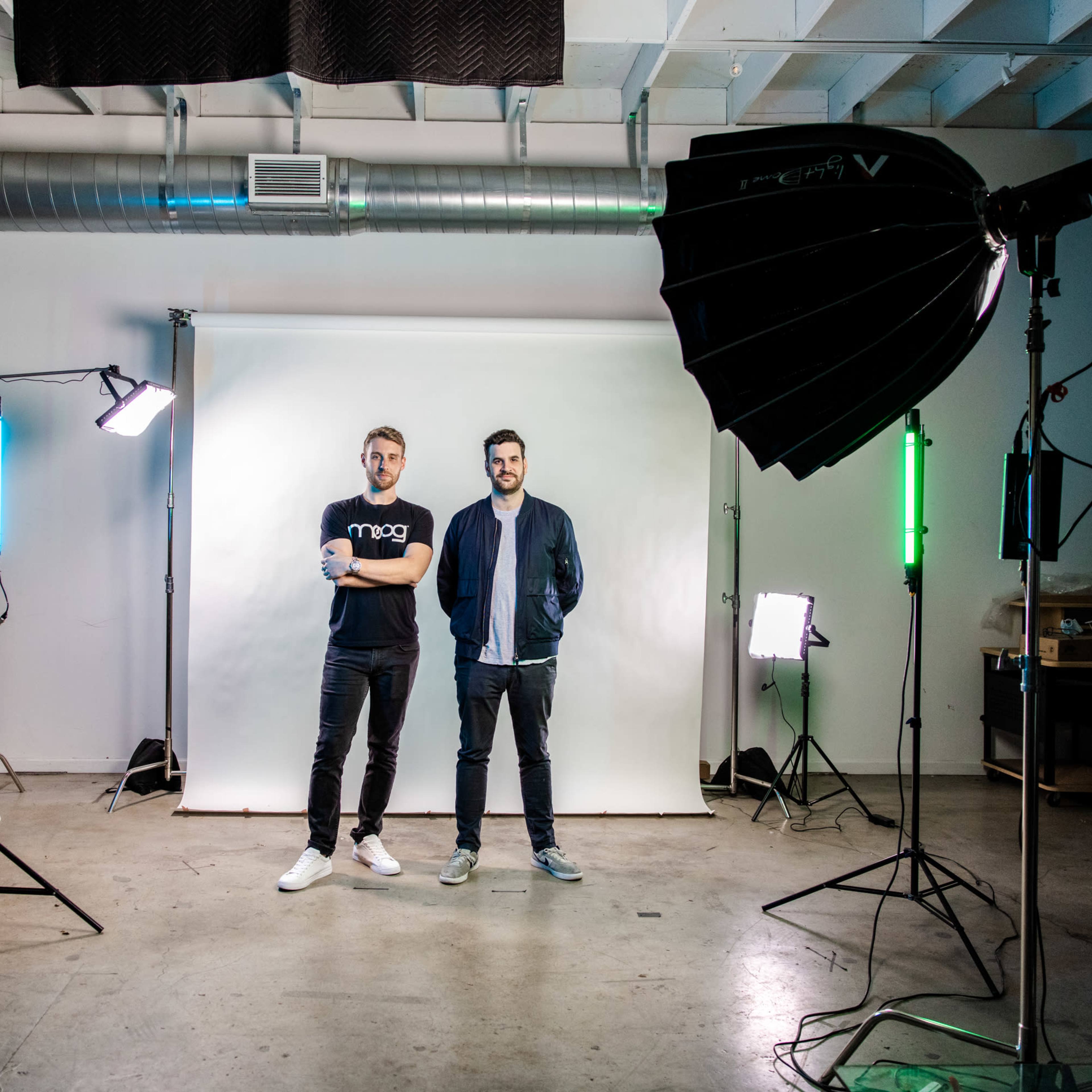 Two men stand in a photography studio, flanked by various lighting equipment and a plain backdrop.