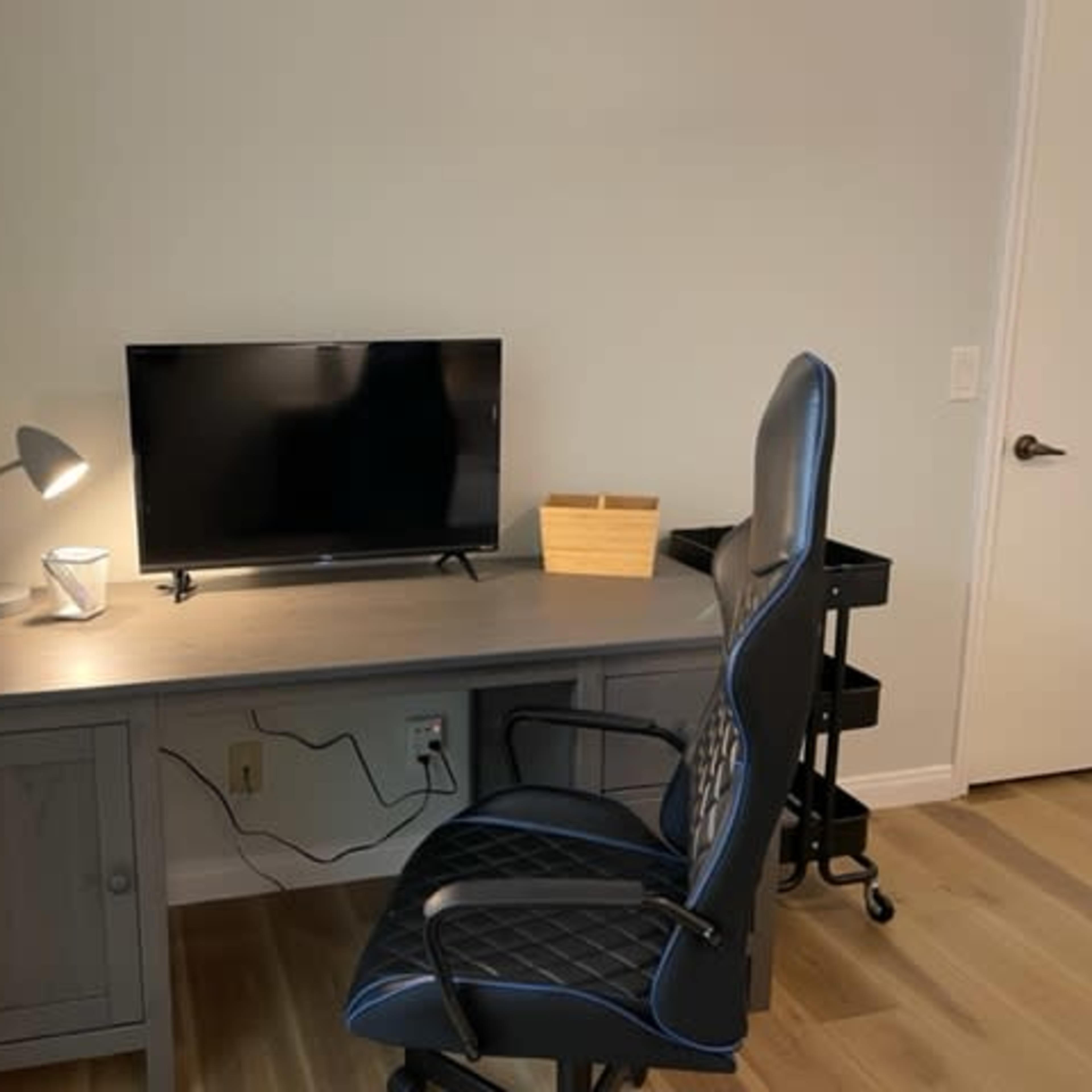 A gray desk with a television on it, a lamp, a small basket, and a black office chair are set against a light-colored wall.