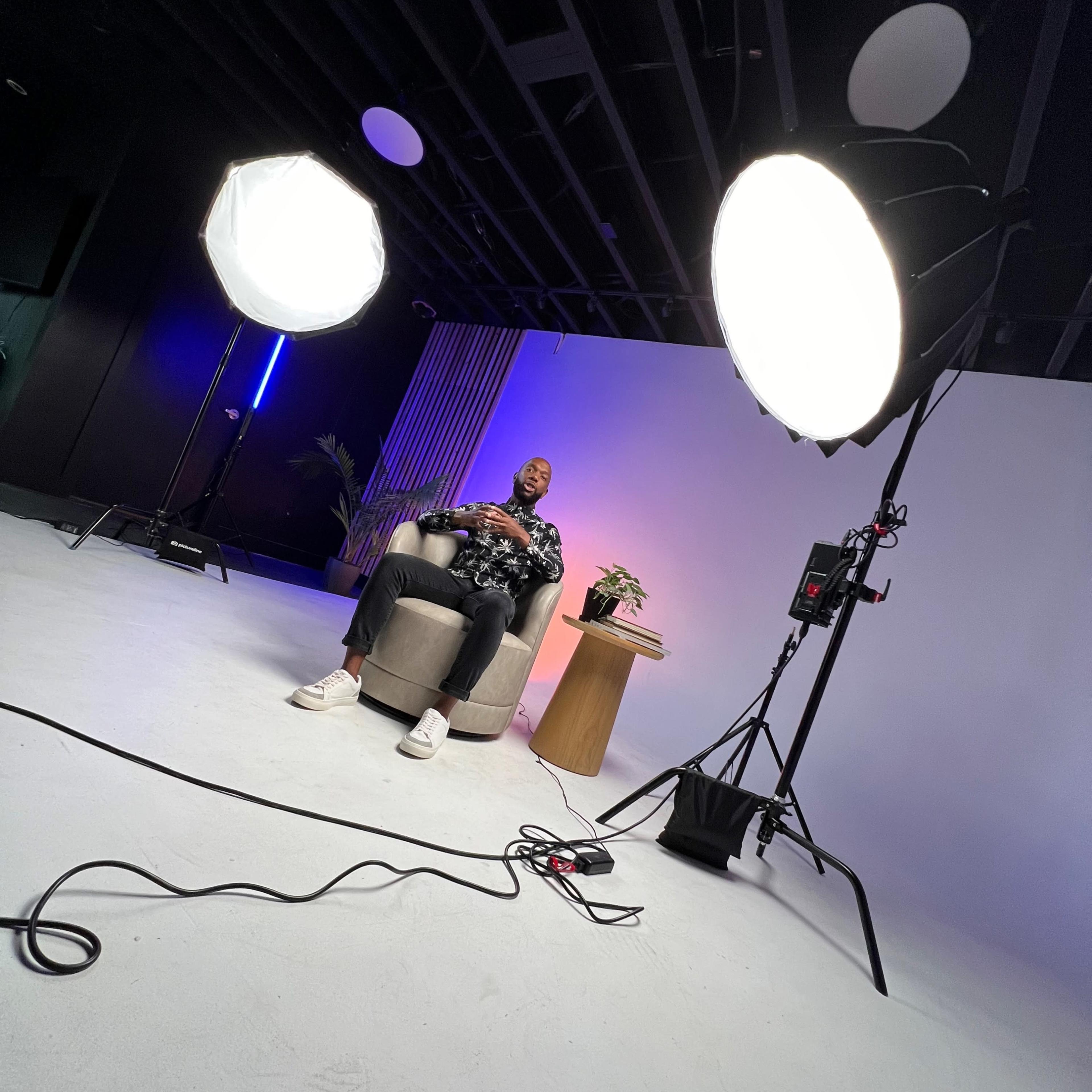 A person sits in a chair in a studio setting, flanked by two large lights and a small table with a potted plant.