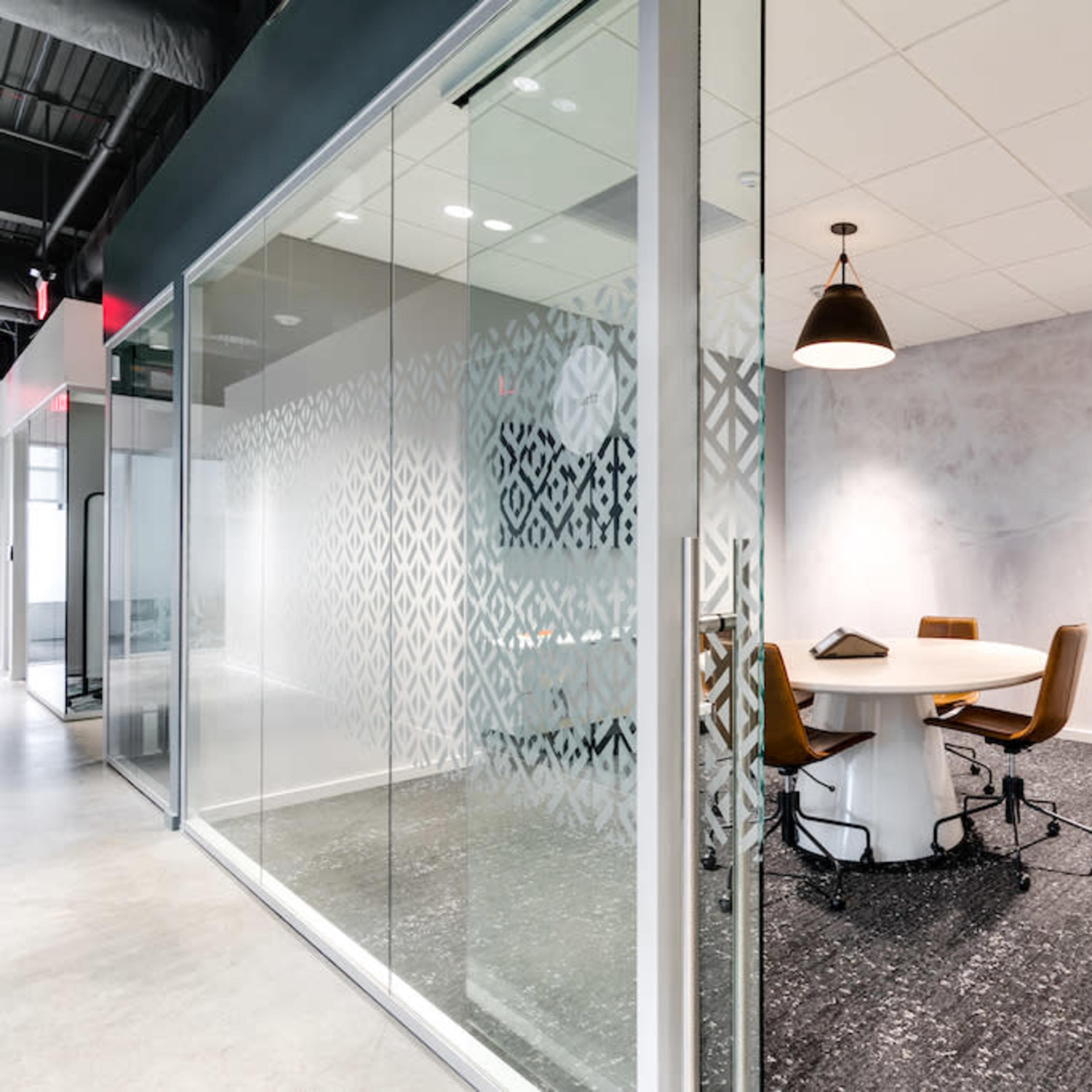 A modern meeting room with a round table and four chairs, visible through a glass wall, alongside a digital control panel mounted on the wall.