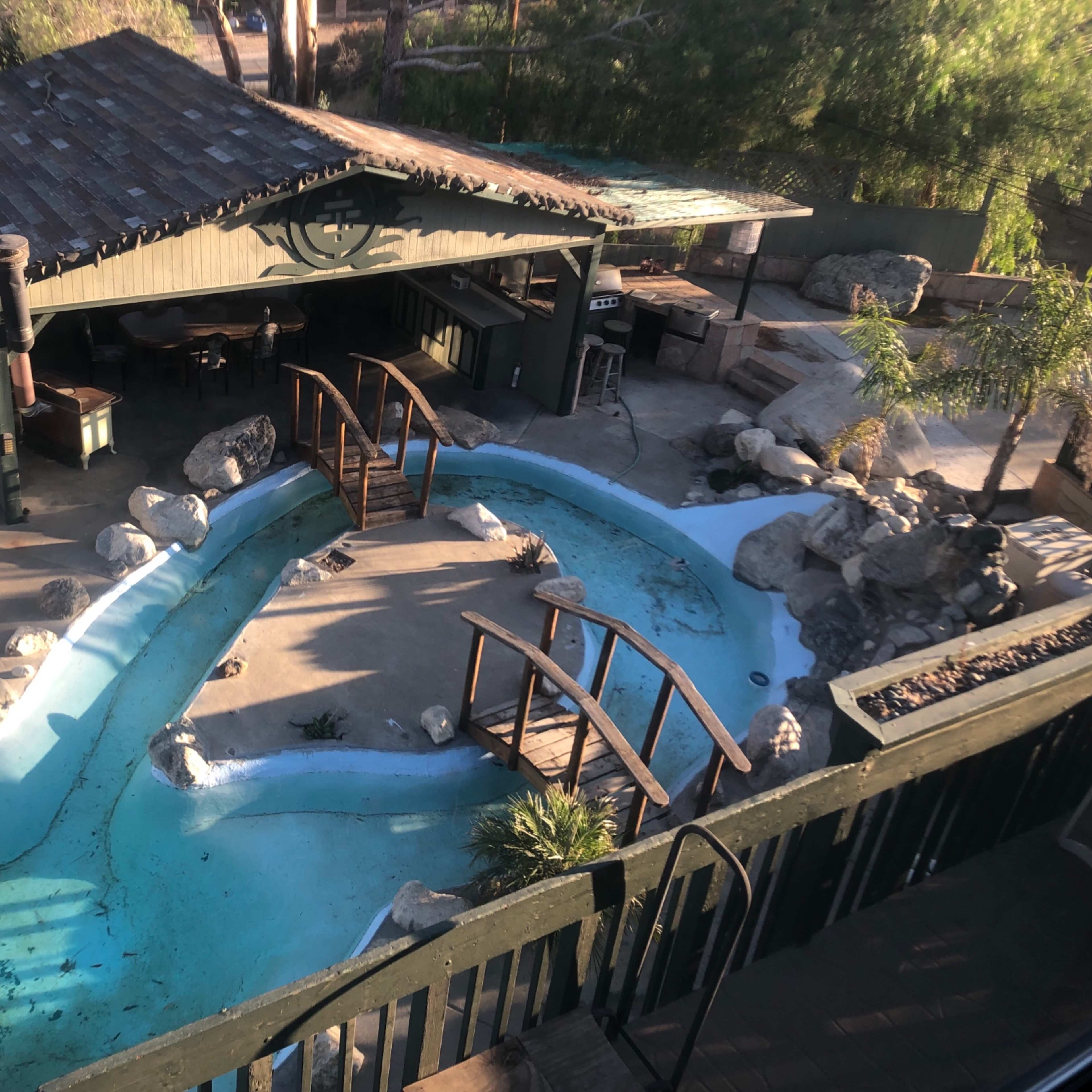A curved pool area surrounded by rocks and a wooden bridge, with a shaded seating area nearby.