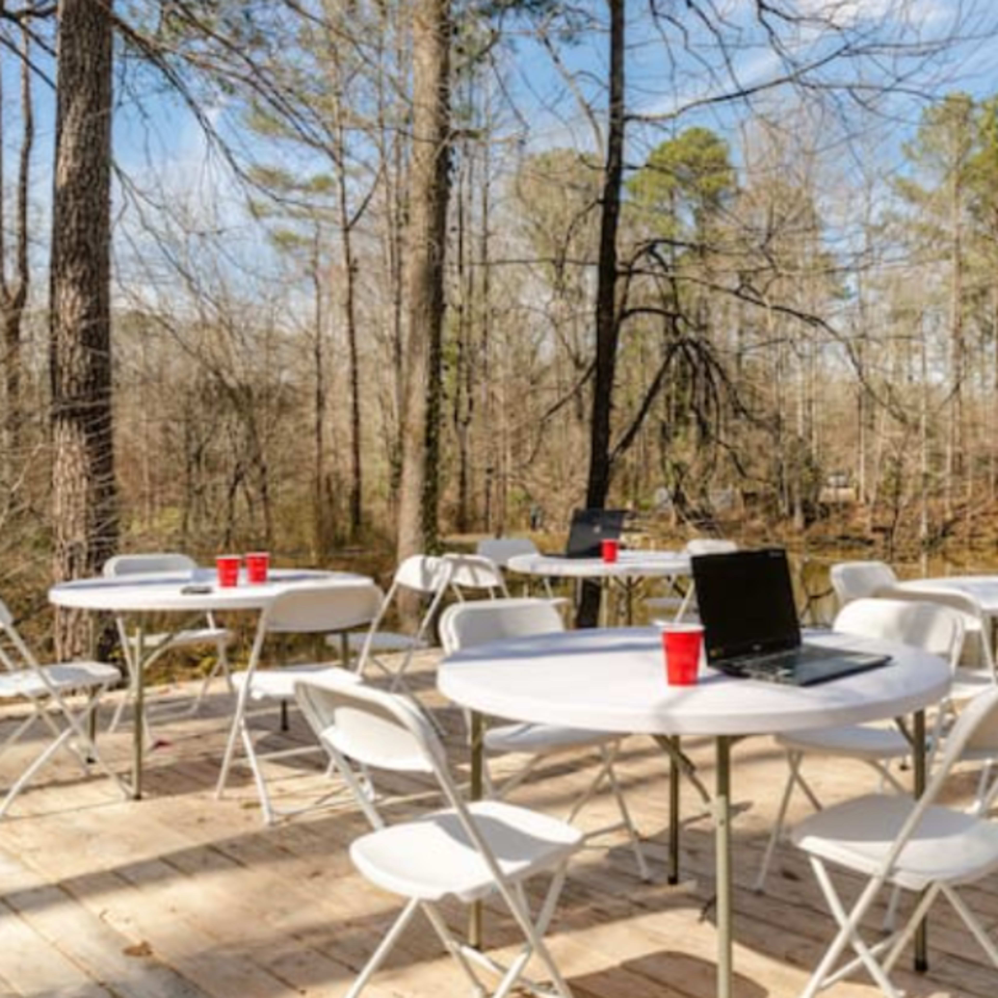 A wooden deck beside a pond is furnished with several white folding tables and chairs, along with a laptop and red cups.