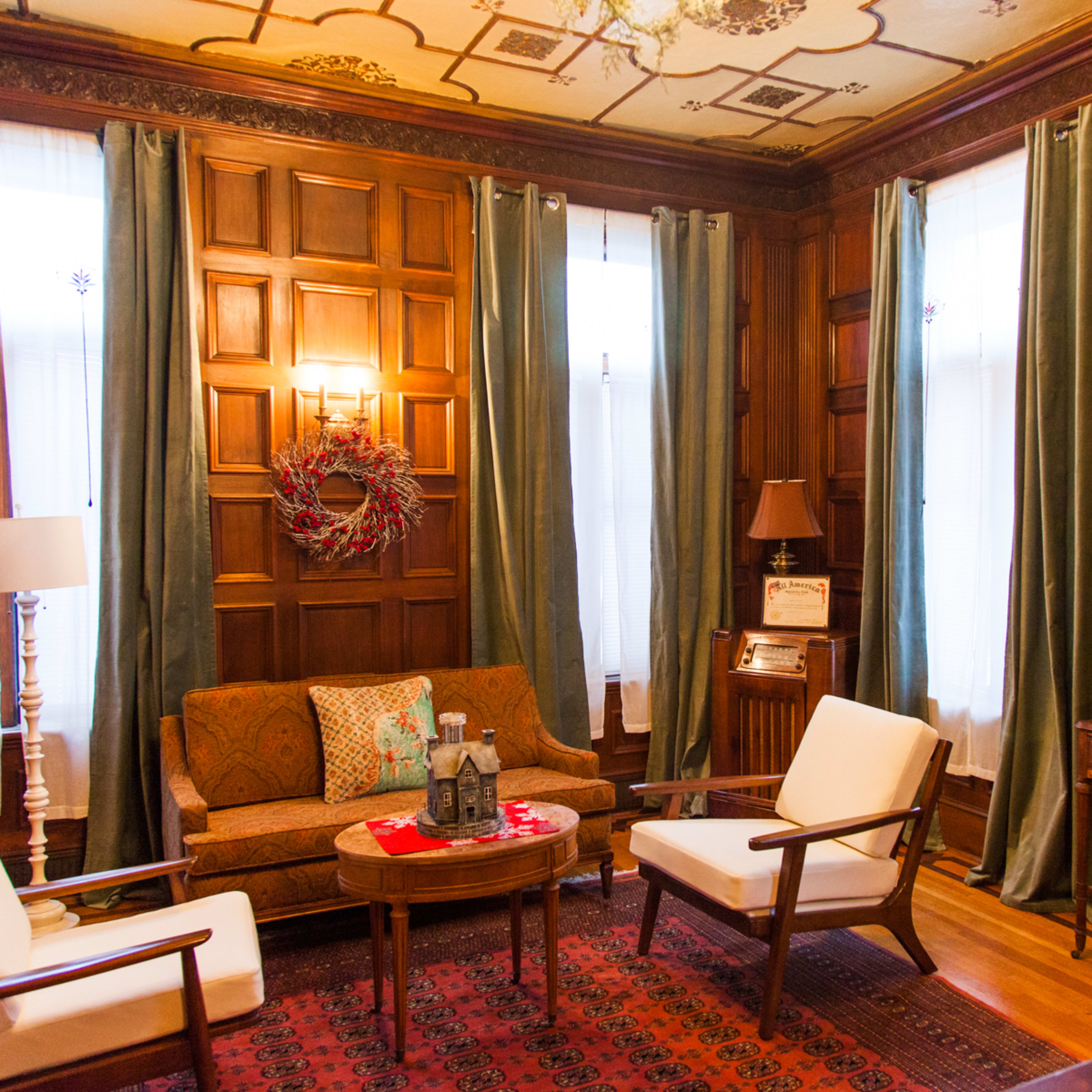 A cozy living room with wooden paneled walls, a sofa, two white chairs, a side table, and a vintage clock on the wall.