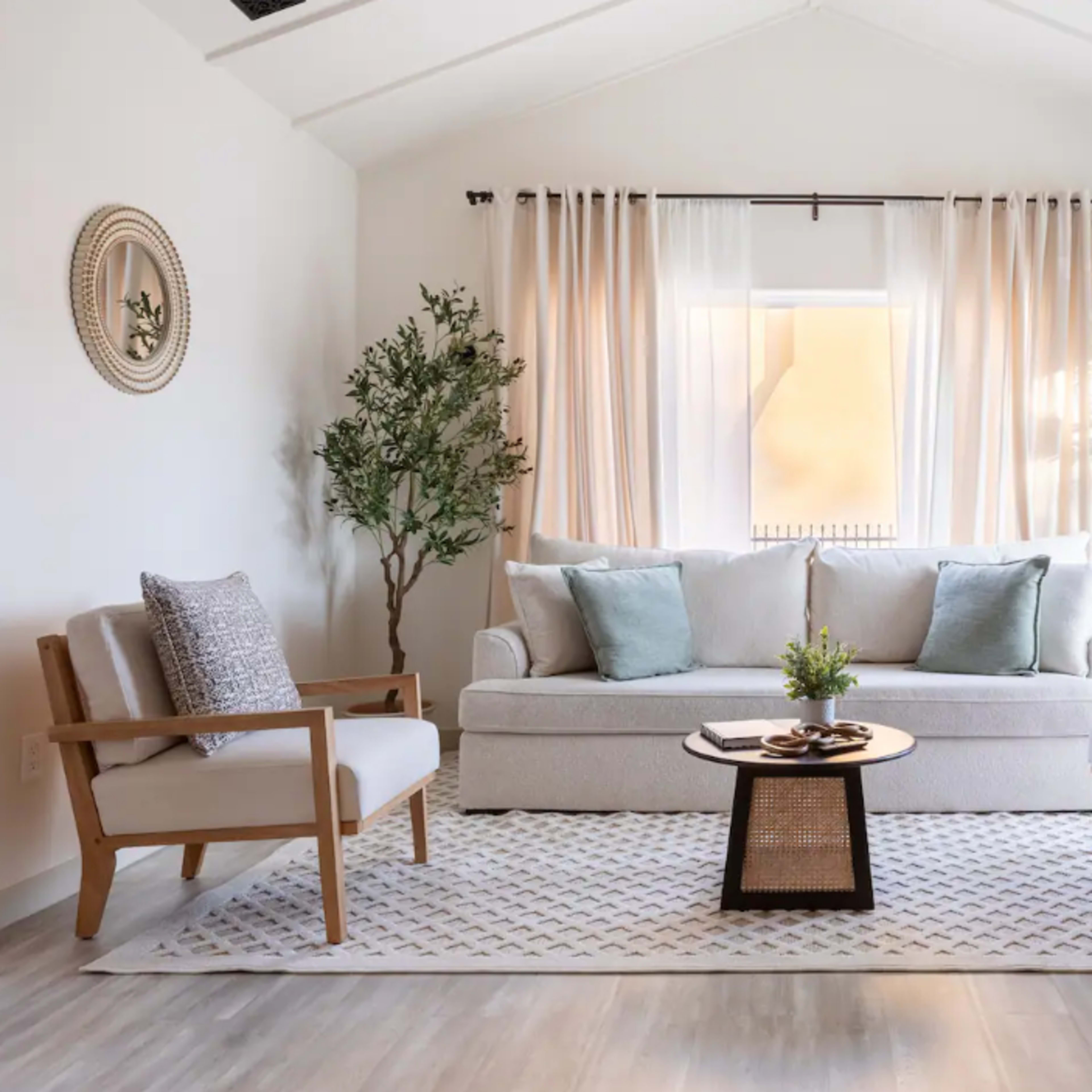 A light and airy living room features a sofa, two armchairs, a round coffee table, and large windows dressed with sheer curtains.