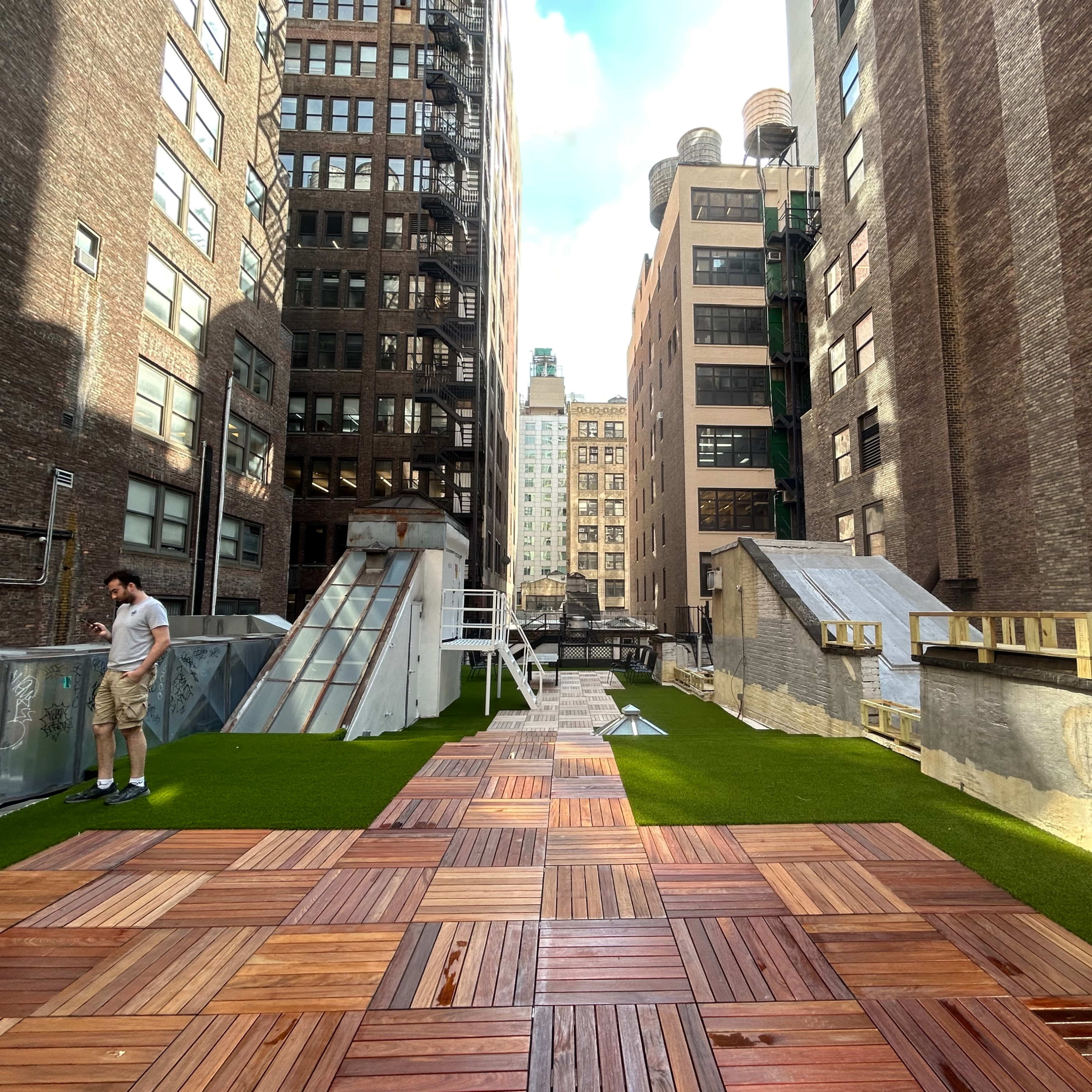A rooftop area with a wooden deck, artificial grass, and various structures, including a small stairway and a chimney, surrounded by tall buildings.
