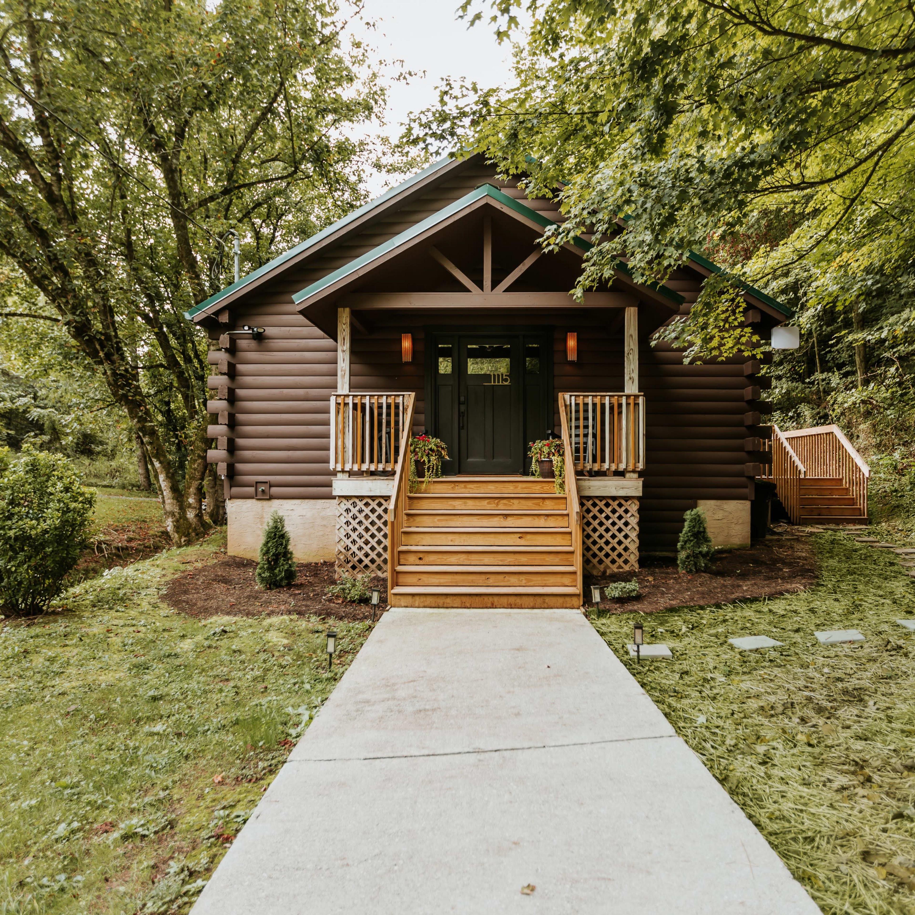 A log cabin with a front porch and wooden steps is set among greenery, leading to a path.