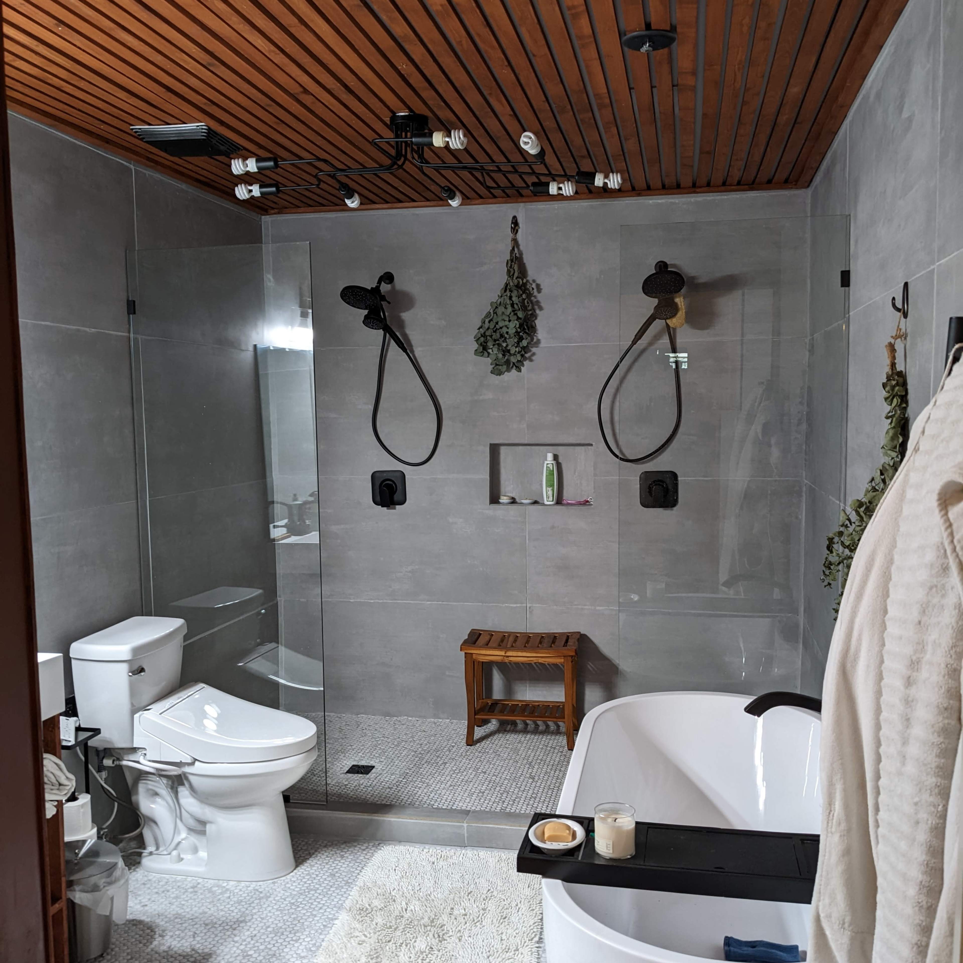 A modern bathroom features a glass shower, a freestanding bathtub, a wooden bench, and a combination of gray tiles and wooden ceiling.