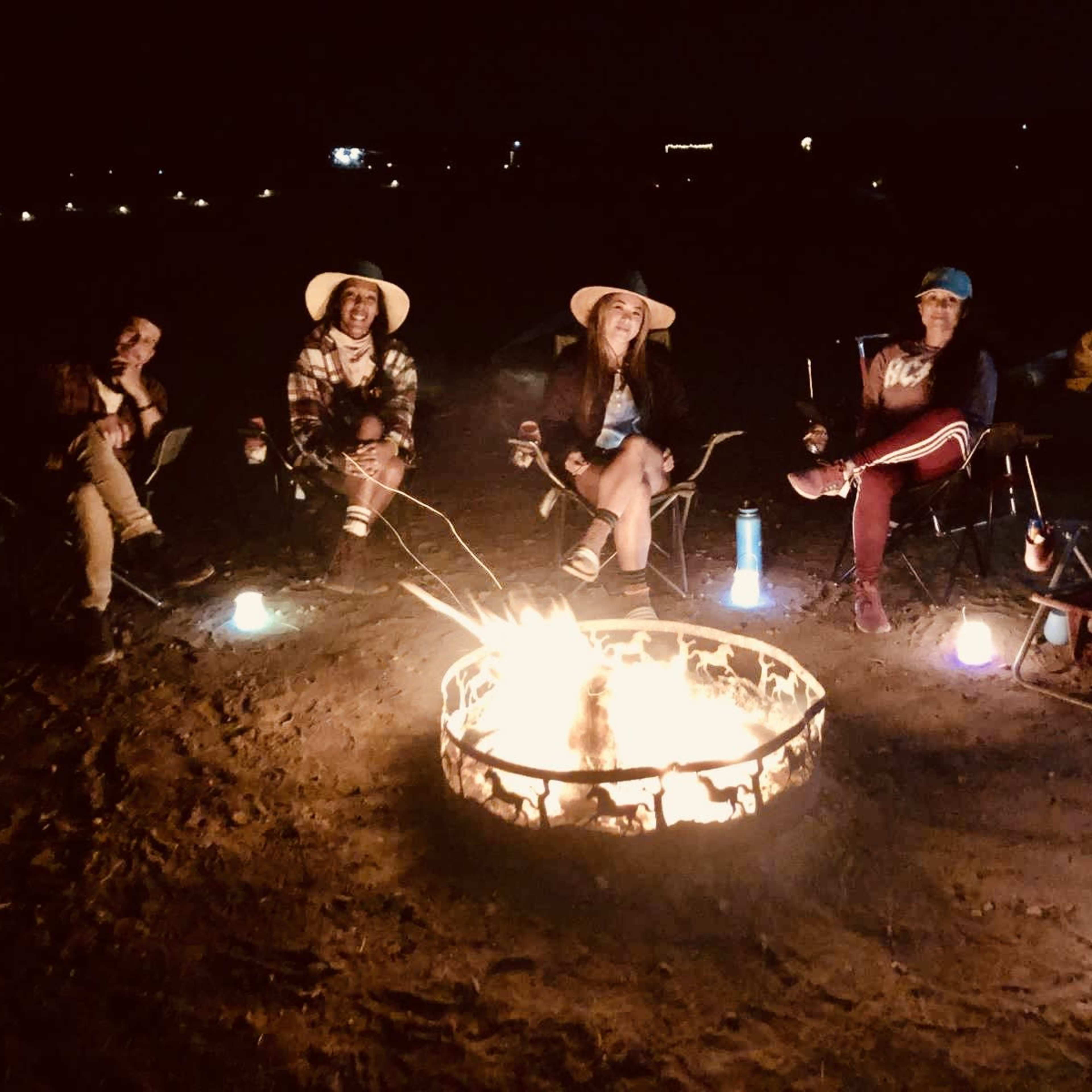 Four people sit around a campfire at night, with camping chairs and glowing lanterns nearby.