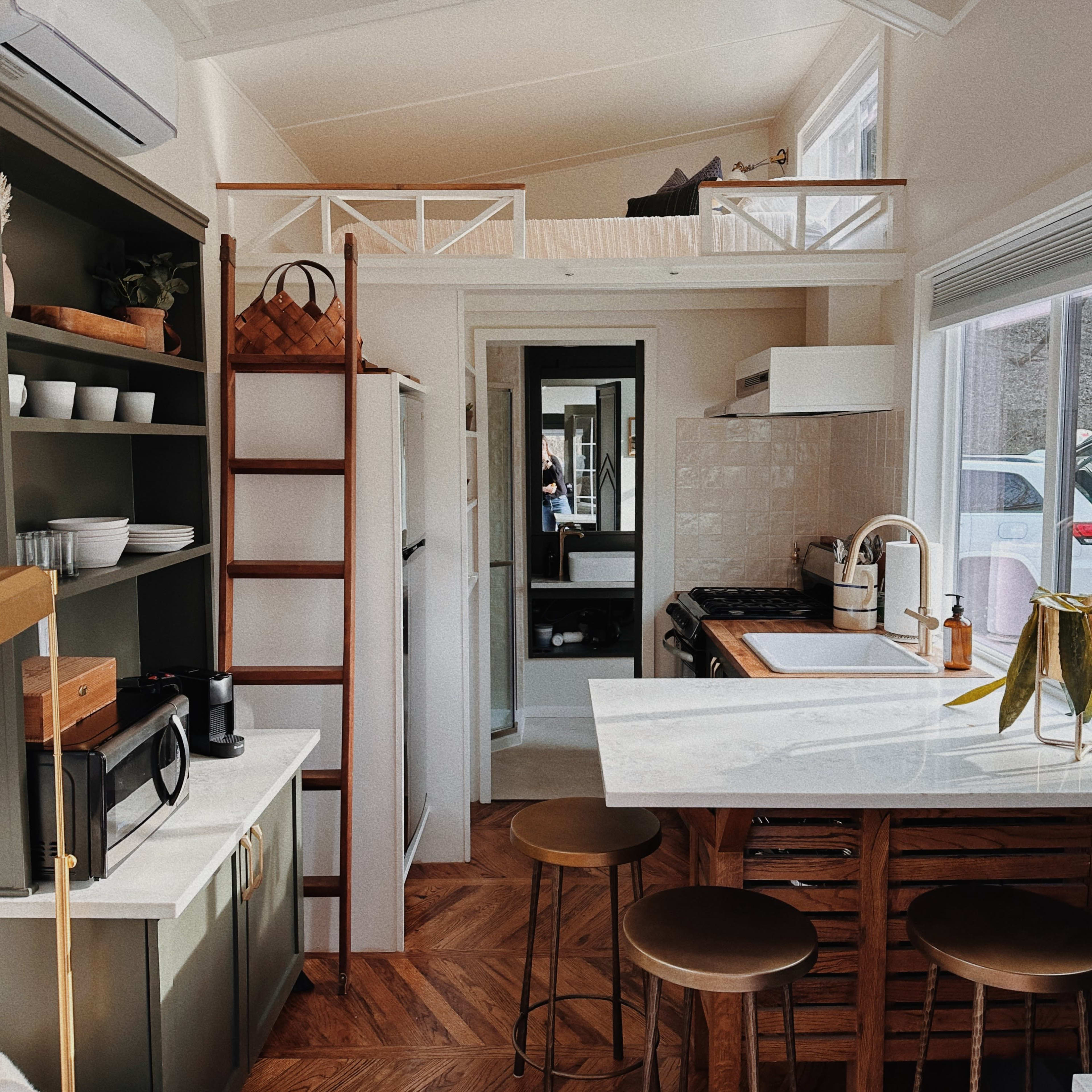 A modern kitchen features a wooden ladder leading to a loft area, with a dining table and bar stools, surrounded by cabinetry and appliances.