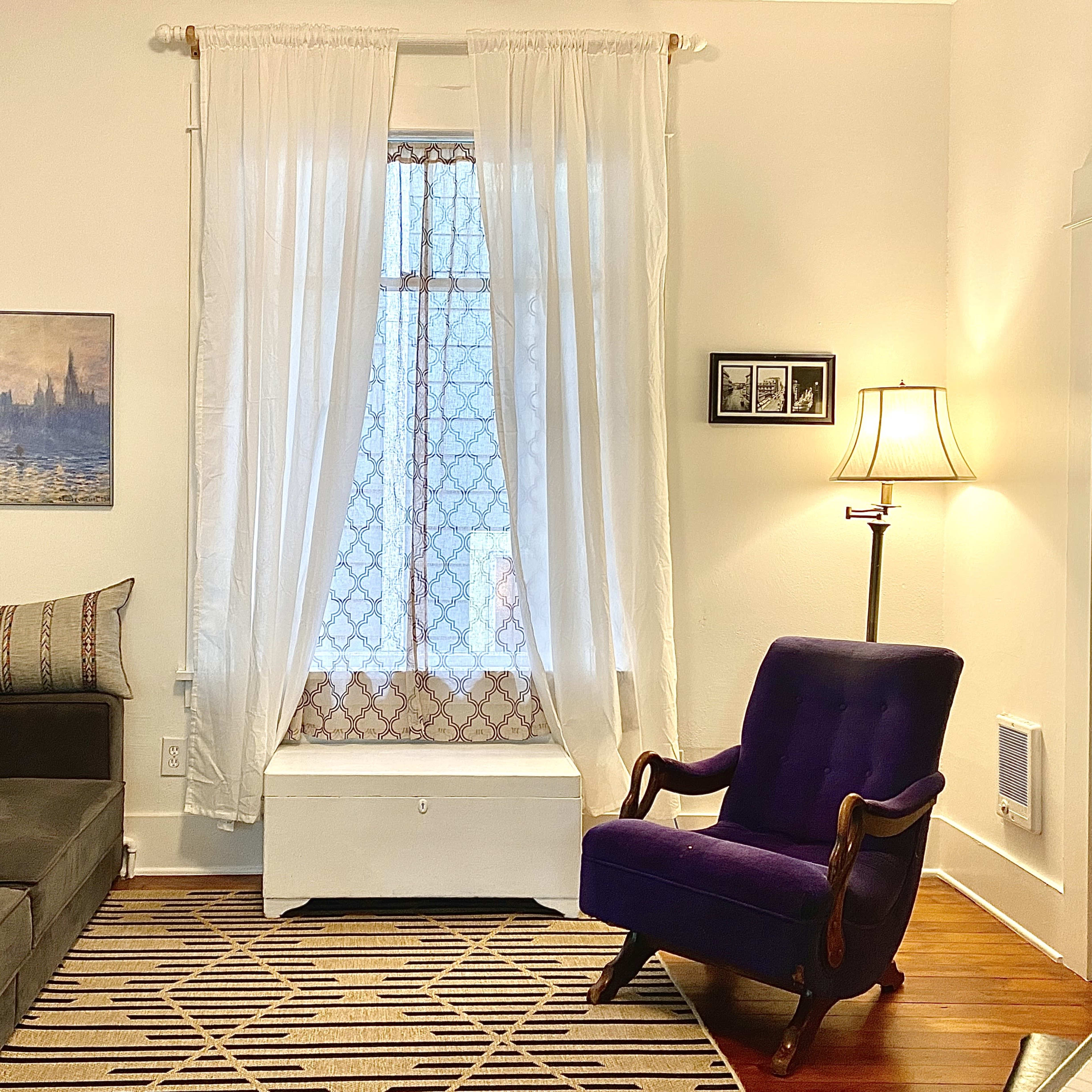 A cozy living room features a gray sofa, a purple armchair, and a window draped with white curtains, illuminated by a table lamp.