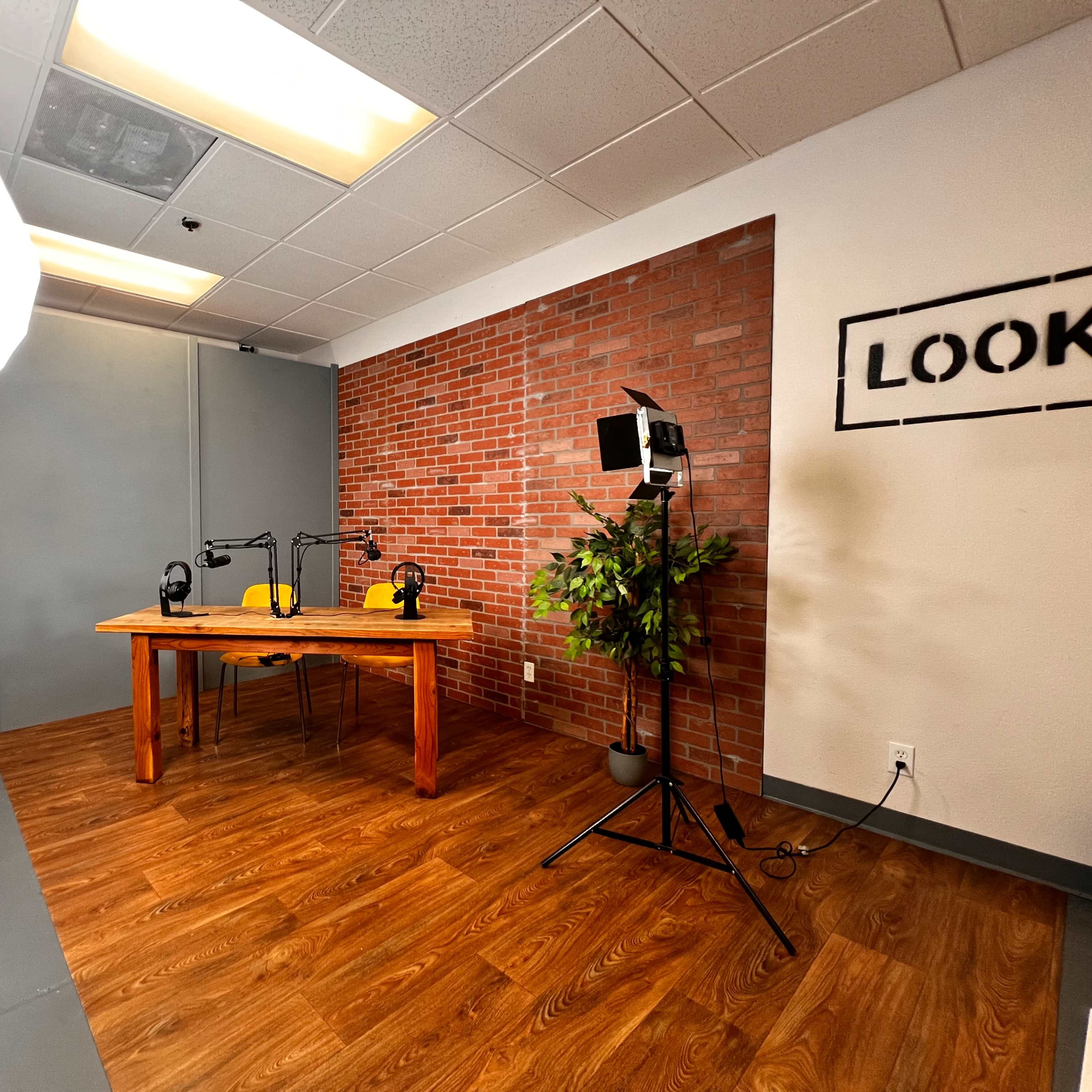 A podcast studio setup with a wooden table, two microphones, and lighting equipment against a brick-patterned wall.